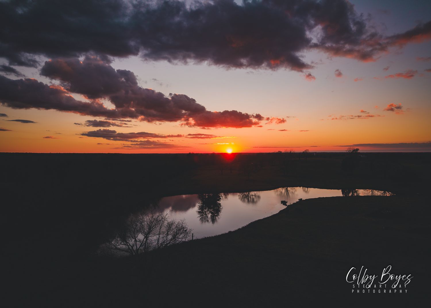 Dramatic sunset silhouettes the landscape as orange light reflects off a calm lake below dark clouds.