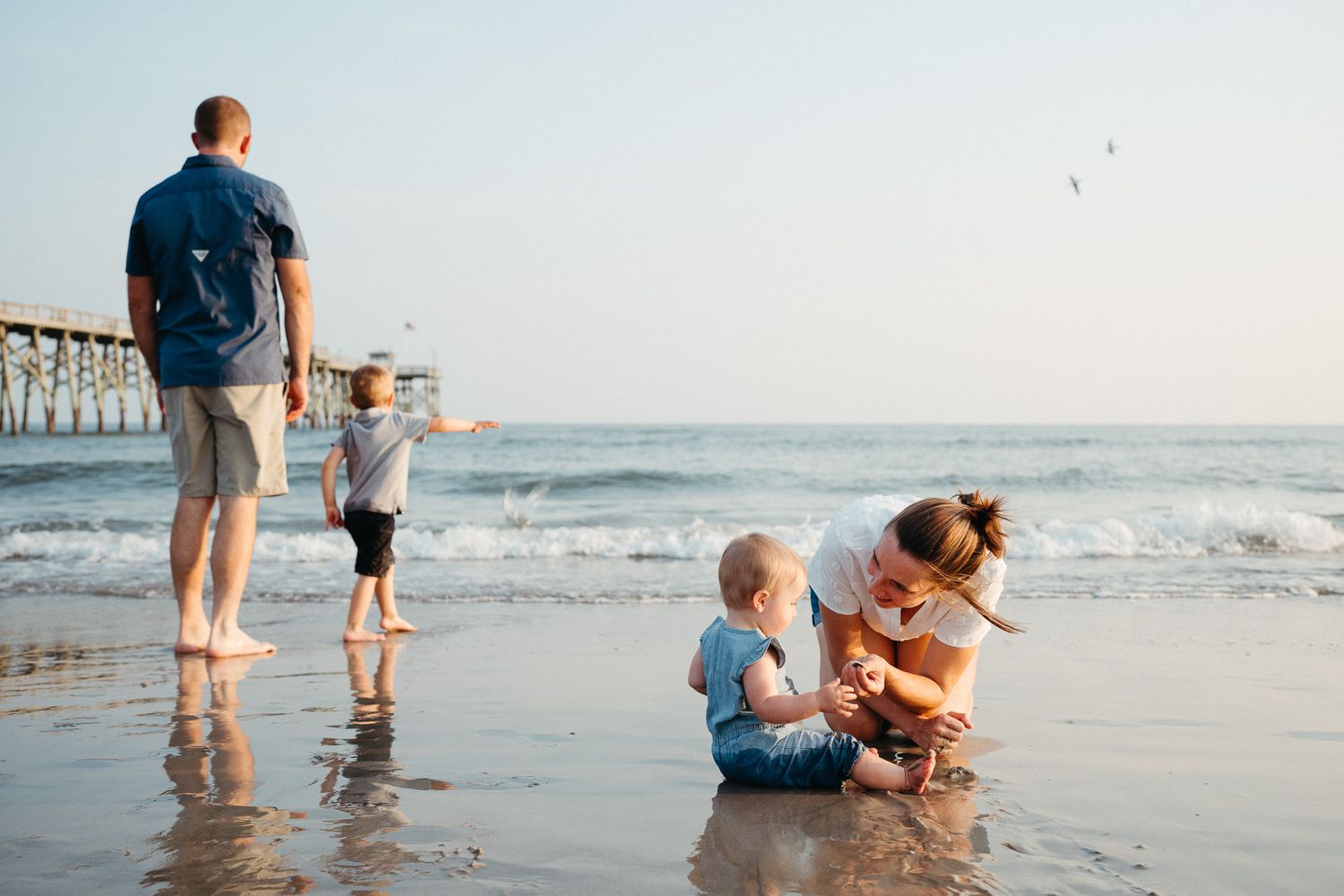 Family enjoying a beach day together near a pier with waves rolling in during sunset. Carolina Beach Family Photography