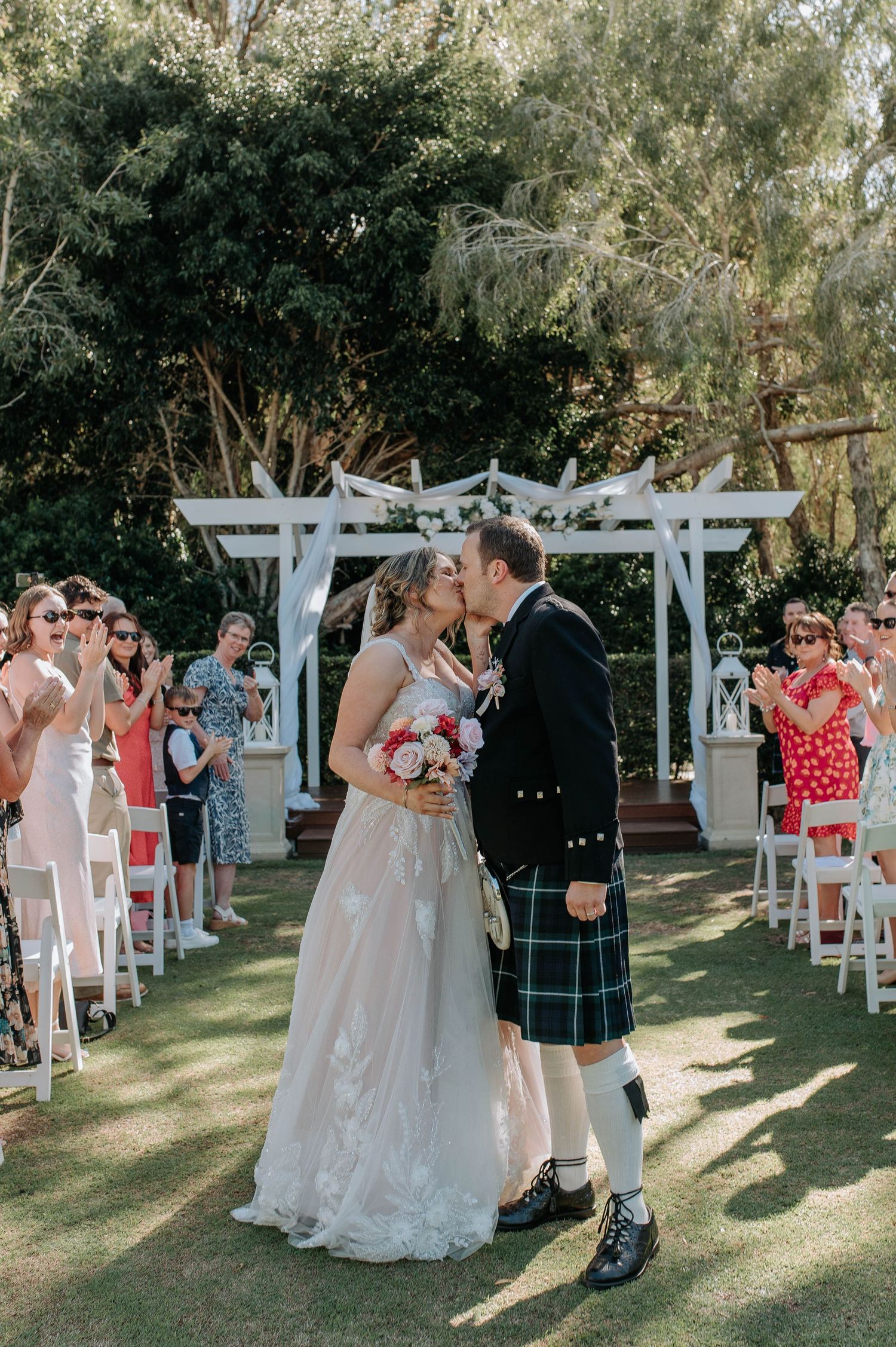 A wedding ceremony takes place under a white pergola with guests watching as the couple shares their first kiss.