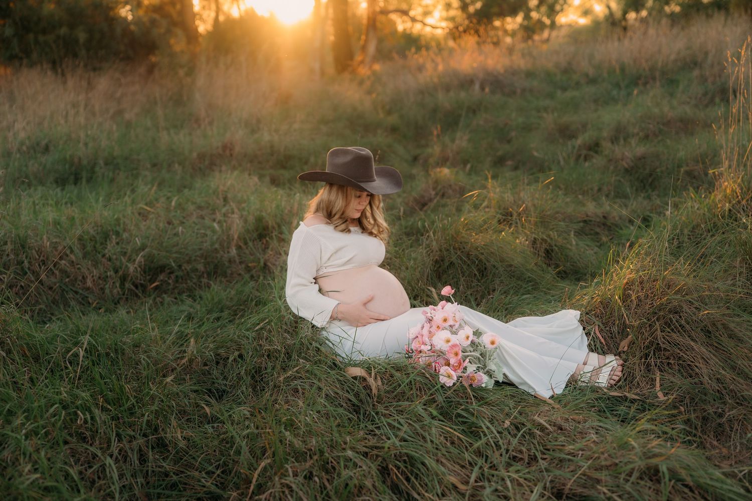 Pregnant woman in white dress and cowboy hat sitting in golden sunset field holding pink flowers.
