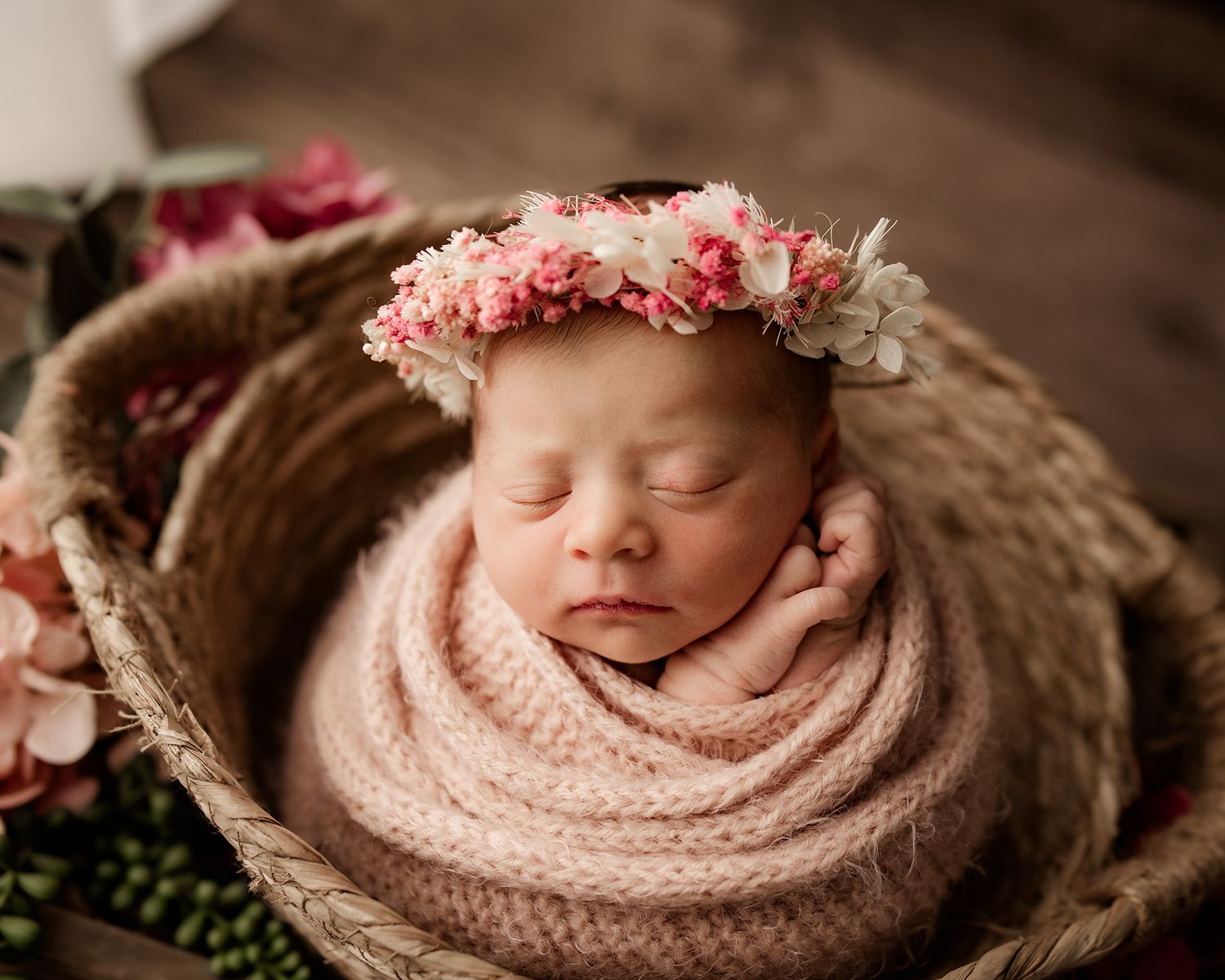 Newborn baby girl sleeping in woven basket, wrapped in pink knit blanket, wearing delicate floral crown.