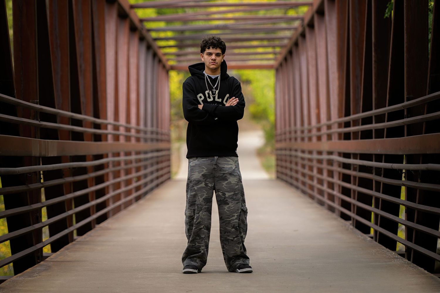 A high school senior in Polo hoodie and camo pants stands arms crossed on a wooden bridge surrounded by red metal railings.