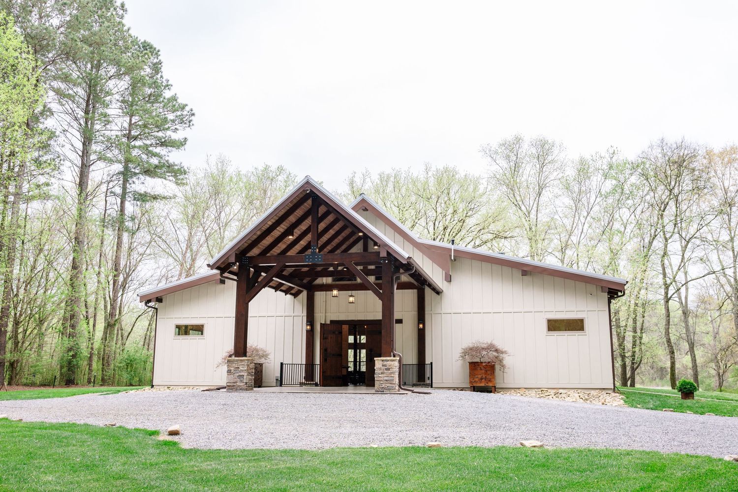 Rustic barn wedding venue with wooden pergola entrance, stone pillars, and lush green trees in spring.