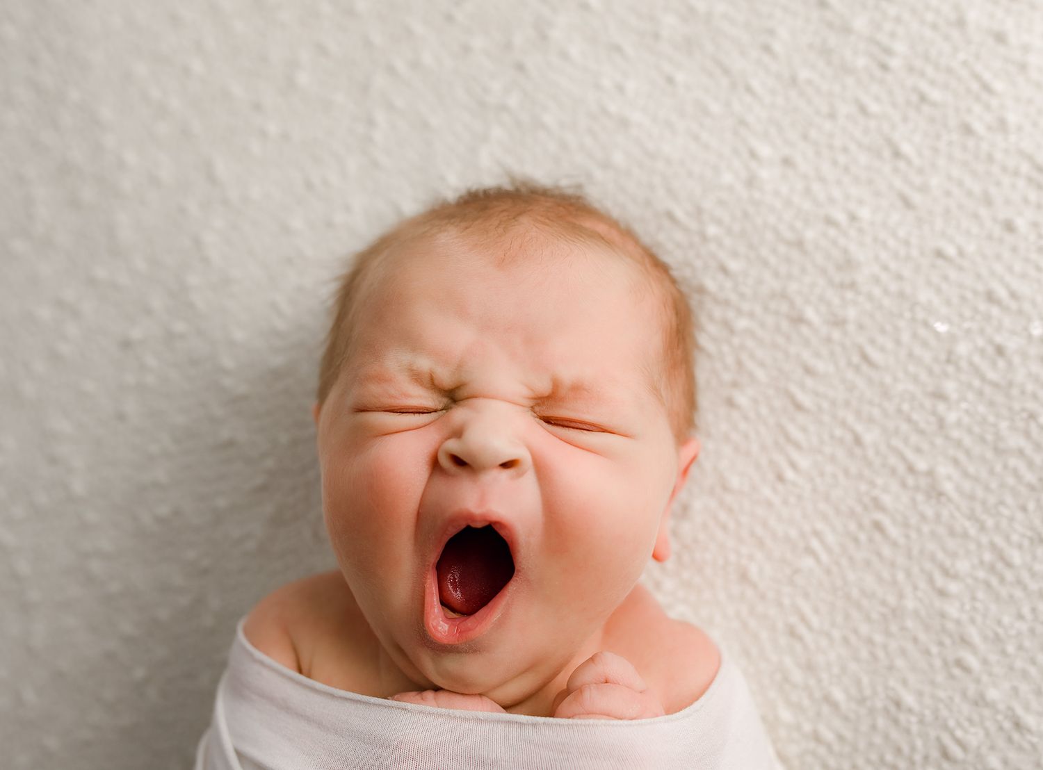 Newborn baby yawning widely while swaddled in white against textured light background.