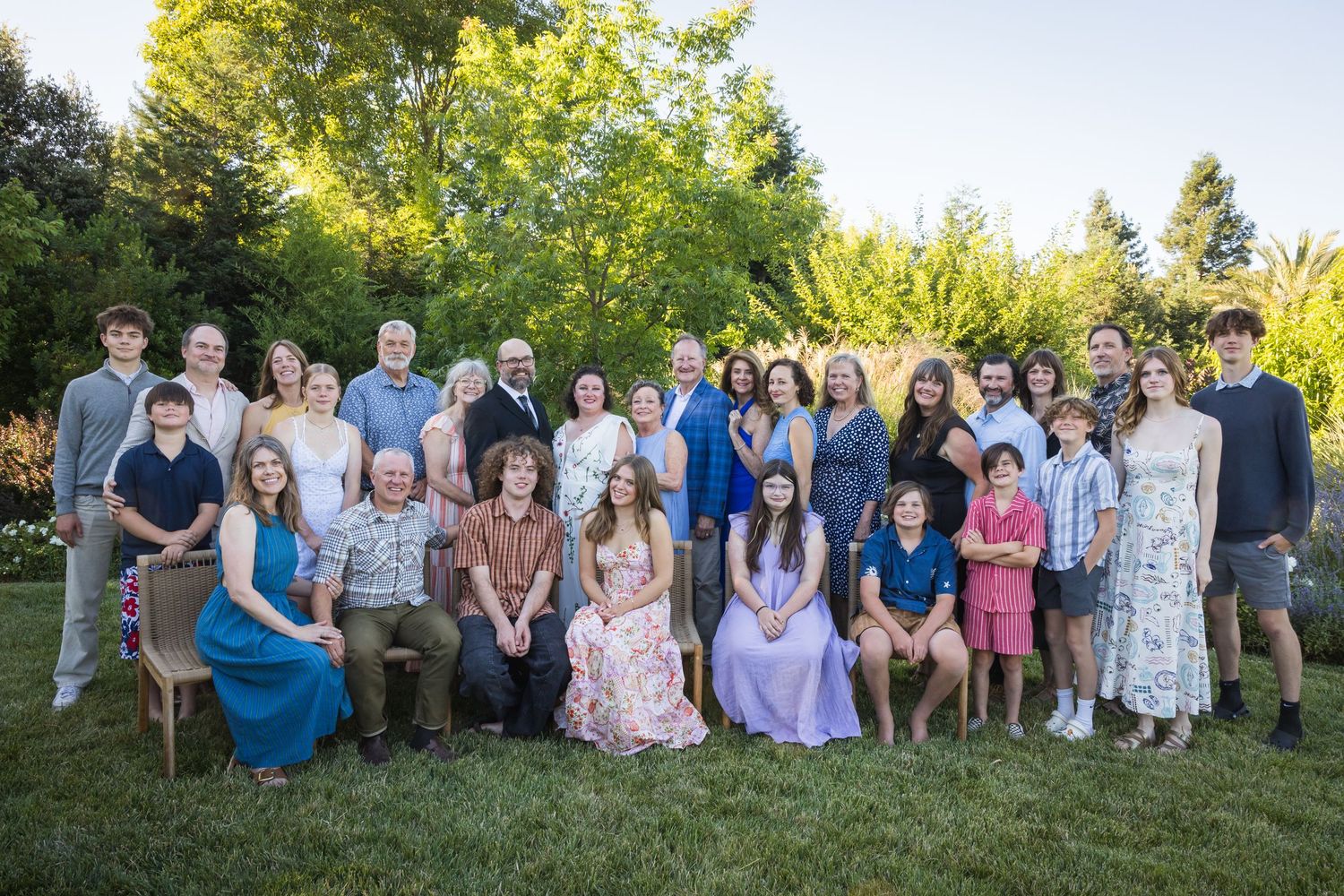 Large multigenerational family group portrait outdoors in lush garden setting, smiling together for formal gathering photo.