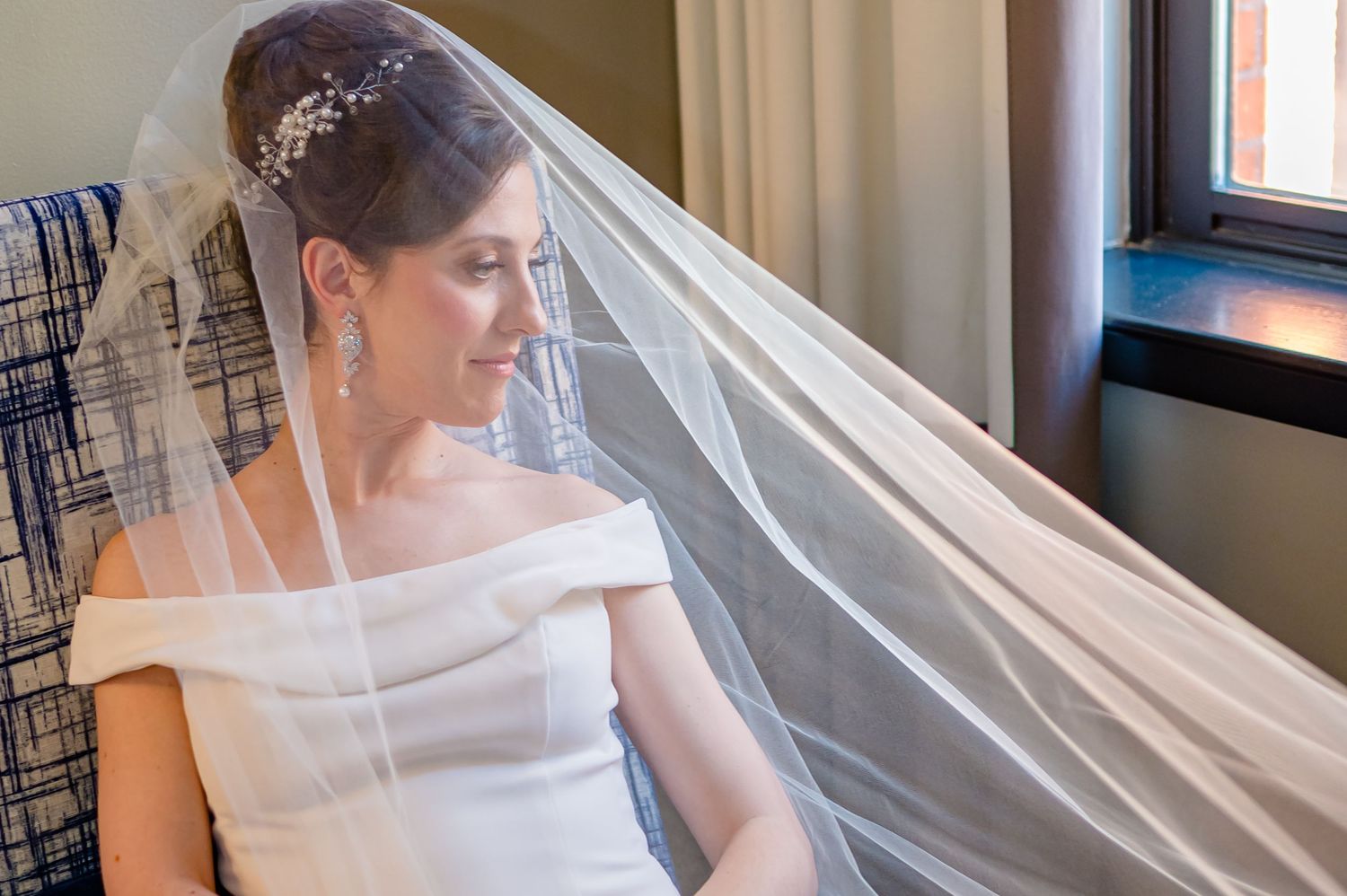 Bride in off-shoulder gown with long veil and hair accessory gazing out window on wedding day.