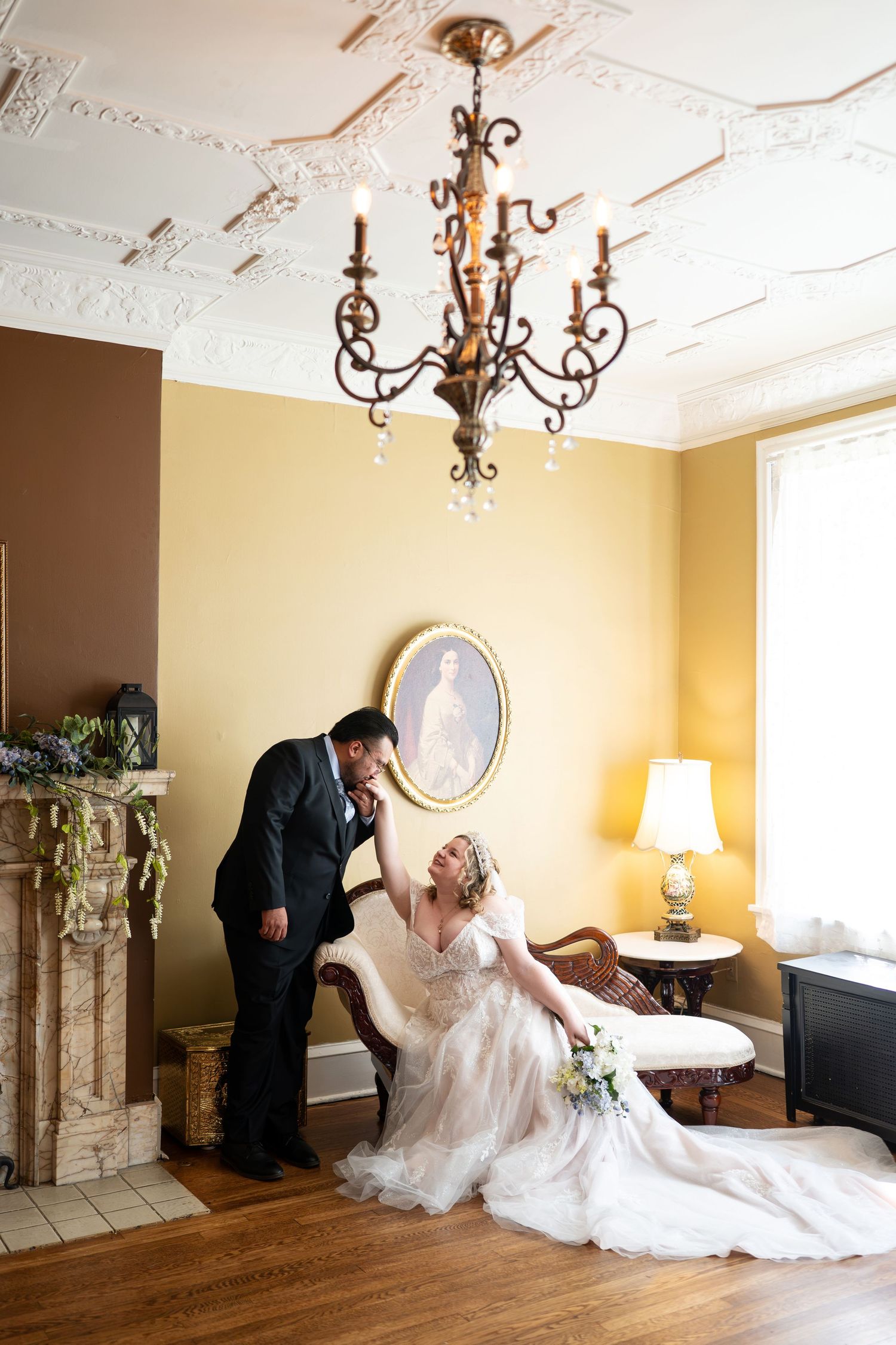 Bride in white gown sits on chaise lounge as groom in tuxedo leans in, under chandelier in elegant yellow room.