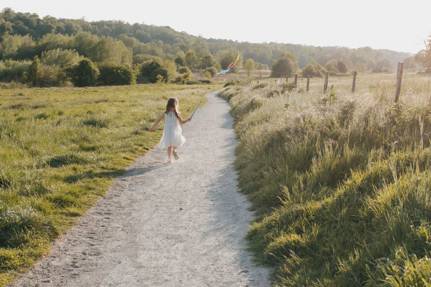 A young girl in a white dress walks along a winding dirt path through a lush green meadow in golden sunlight.