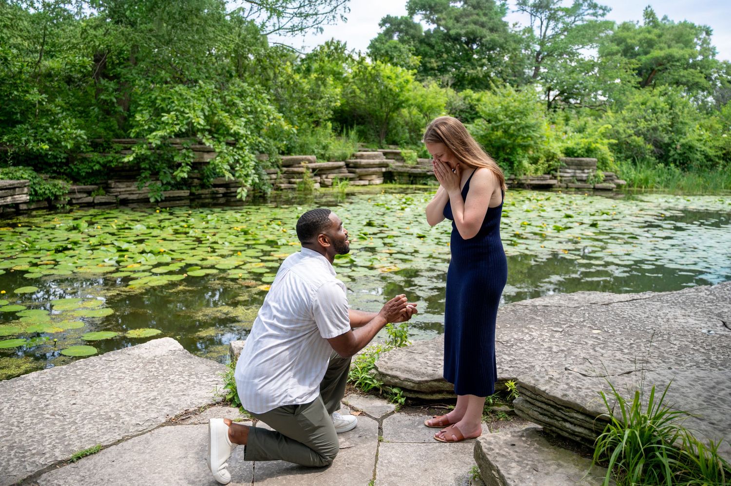 Man proposing to woman on one knee beside a lily pad pond in a lush green garden setting.