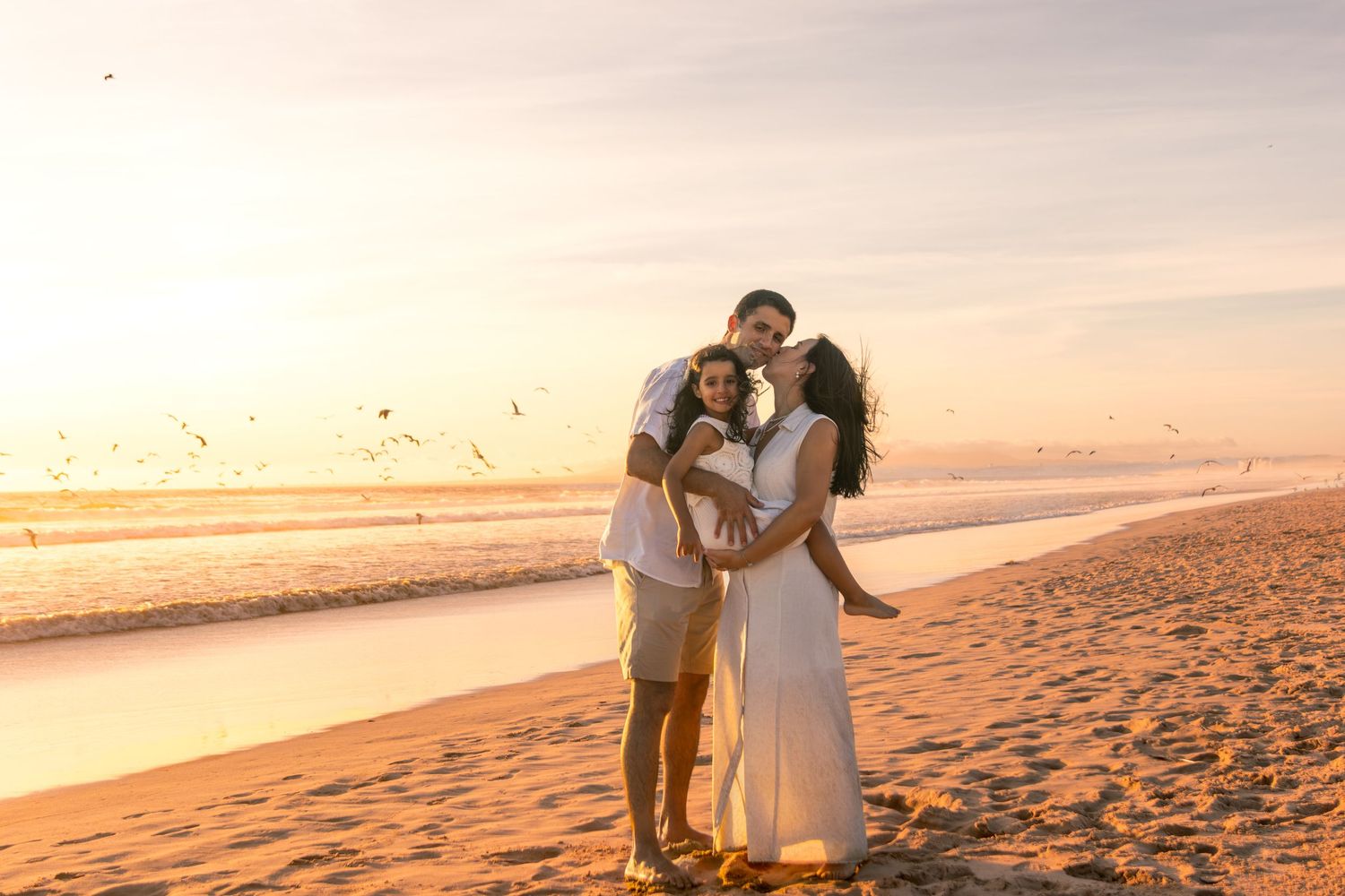 A couple shares a romantic moment on a beach at sunset as seagulls fly overhead.