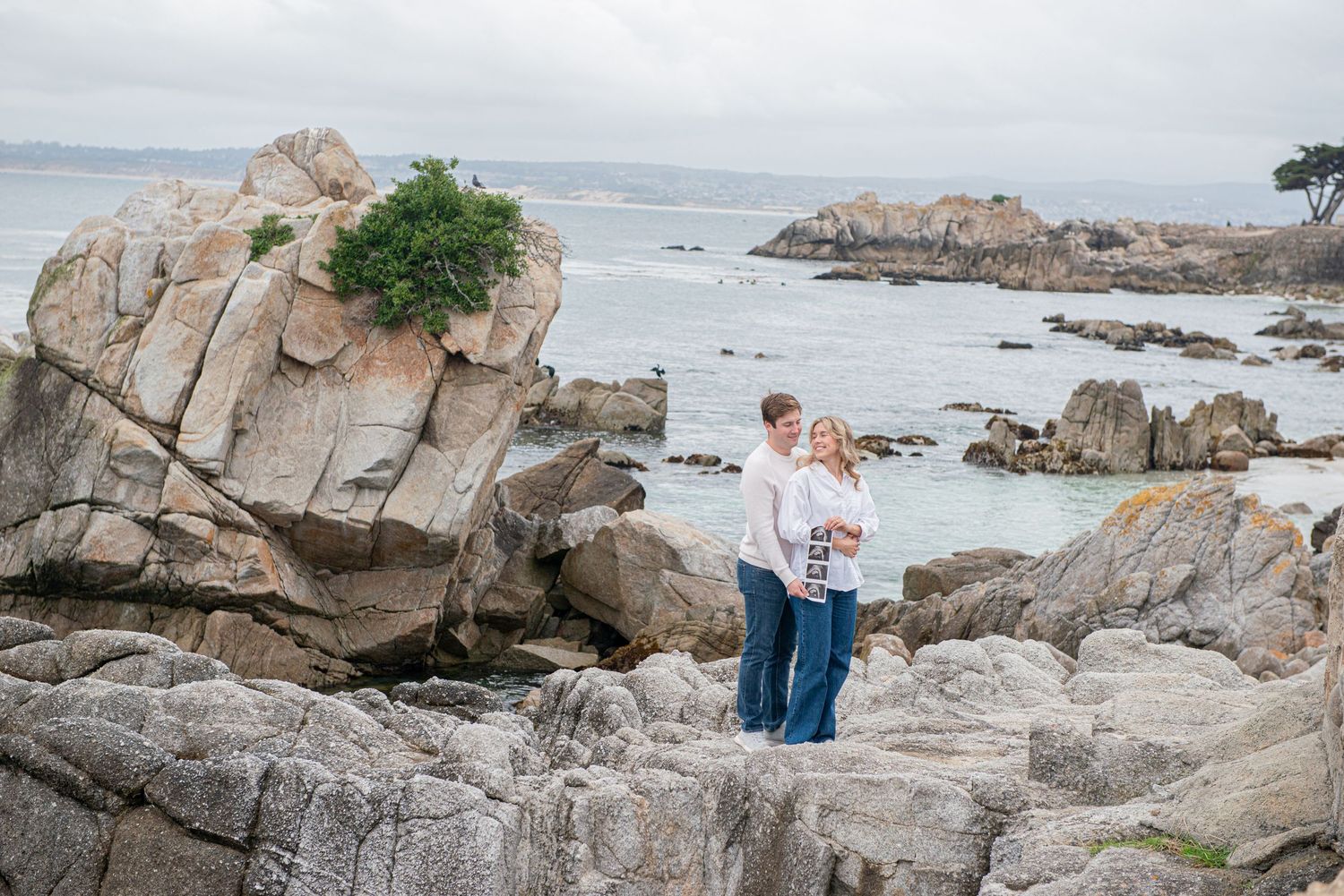 Couple embracing on rocky coastal cliffs with ocean waves and misty sky in background.