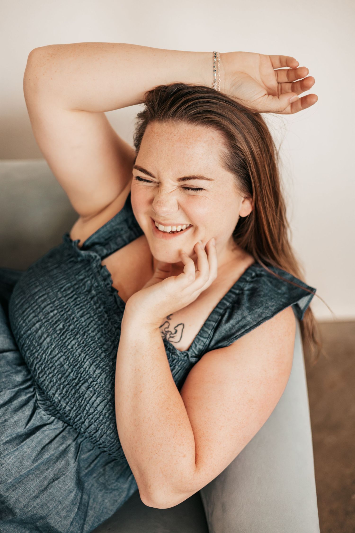 A plus-size woman with freckles laughs joyfully on a sofa, wearing a blue top with a tattoo visible on her arm| Photography Business Coach