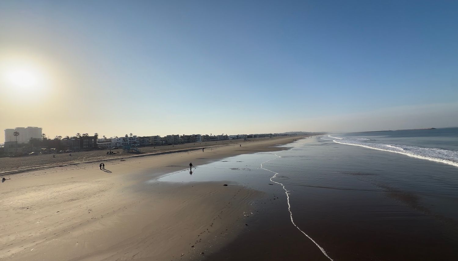 Aerial view of a vast sandy beach at sunset with gentle waves, scattered beachgoers, and a city skyline in the distance.