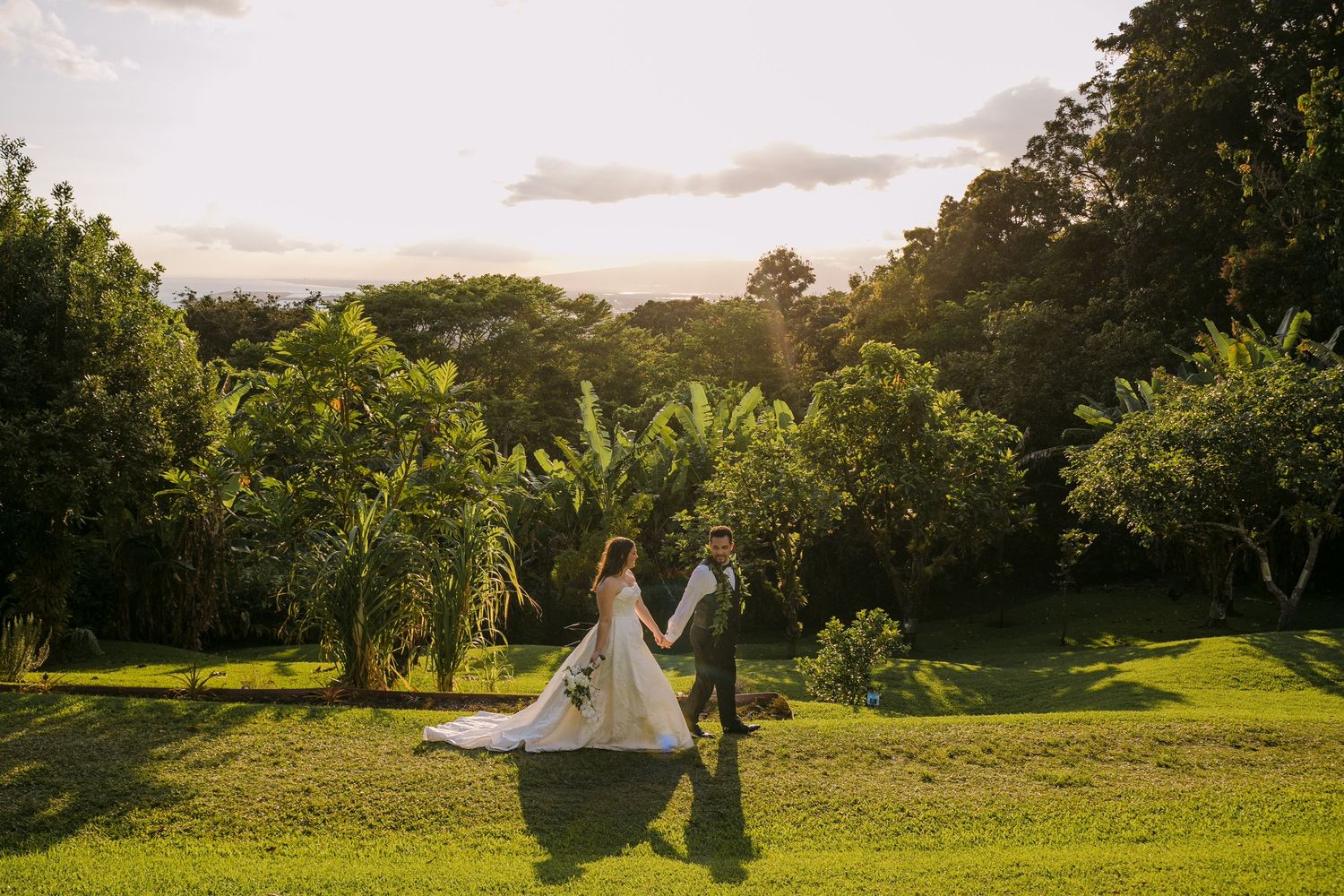 Bride and groom holding hands walking across lush green lawn at sunset with tropical trees in background.