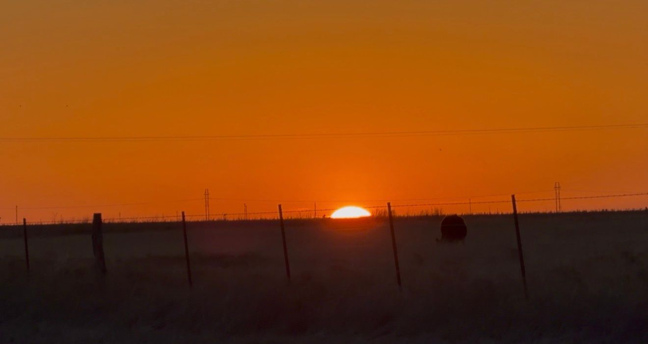 Stunning orange sunset over flat plains with silhouetted fence posts and hay bale on the horizon.