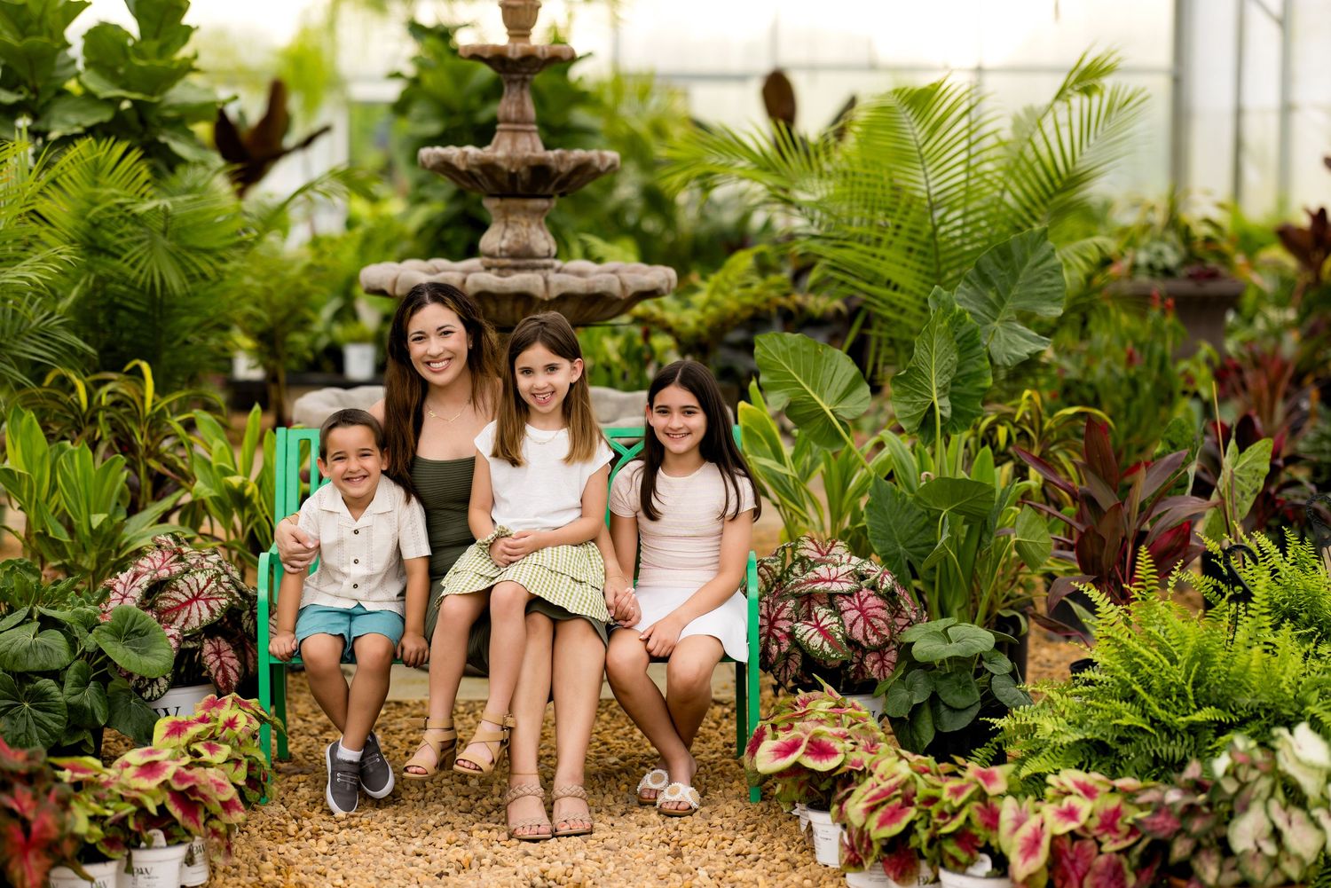 Four girls smiling together on green bench surrounded by lush tropical plants and stone fountain in a greenhouse.