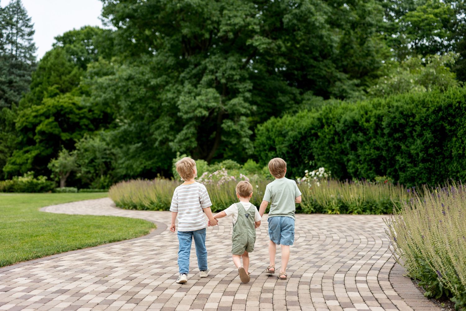 Three young children holding hands walking together on a brick garden path surrounded by lush green trees and plants.