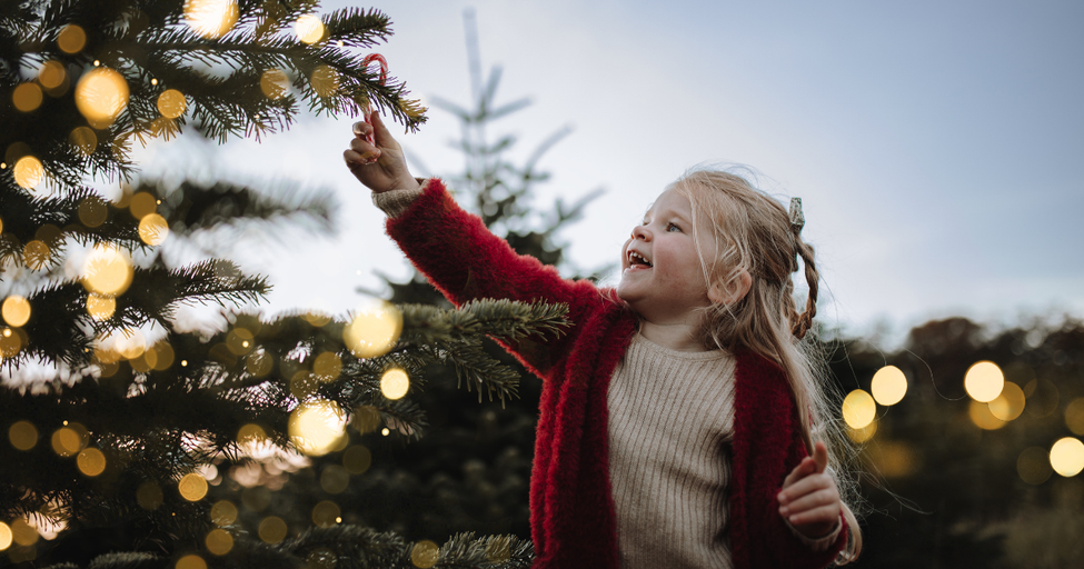 Smiling little girl in a red cardigan reaches up to touch a glowing string light on a Christmas tree outdoors at dusk.