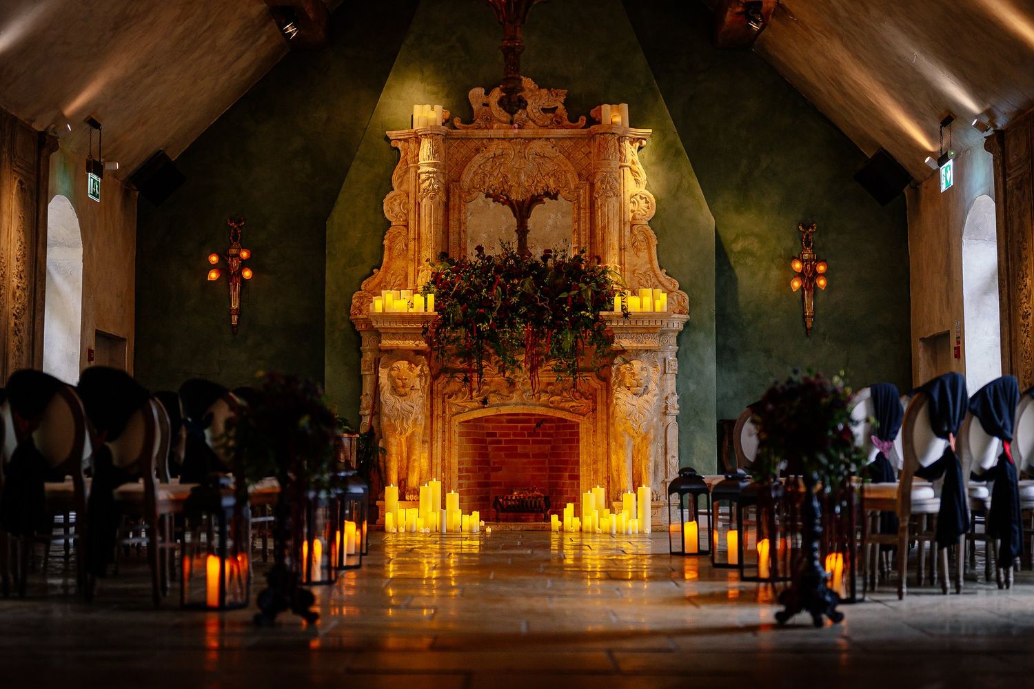 Candlelit wedding ceremony aisle with black chairs leading to an ornate carved stone fireplace adorned with a lush greenery garland.