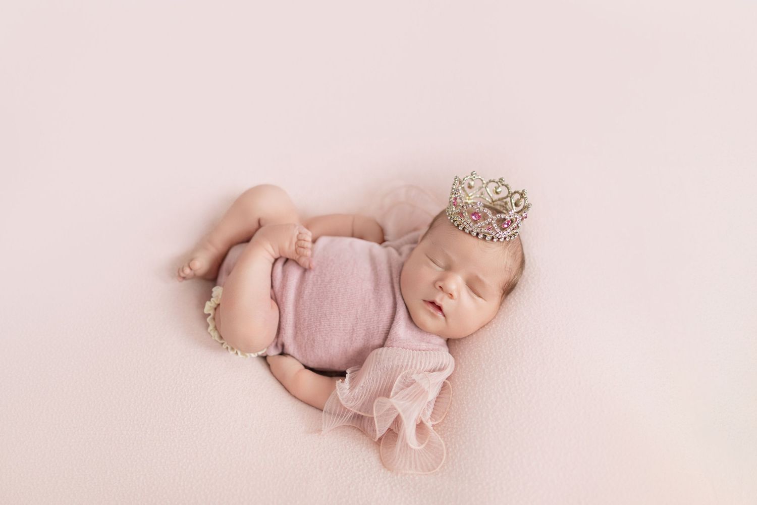 Sleeping newborn baby girl in pink romper and tiny crown on soft pink blanket.