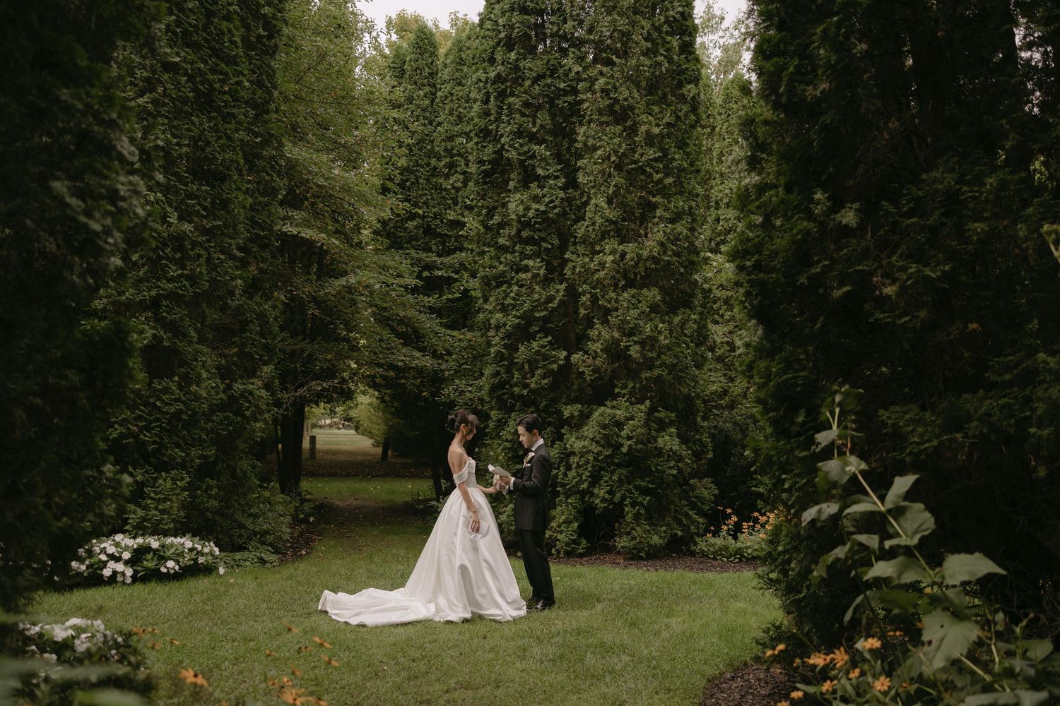 Bride and groom stand face to face surrounded by tall lush green trees in a formal garden.