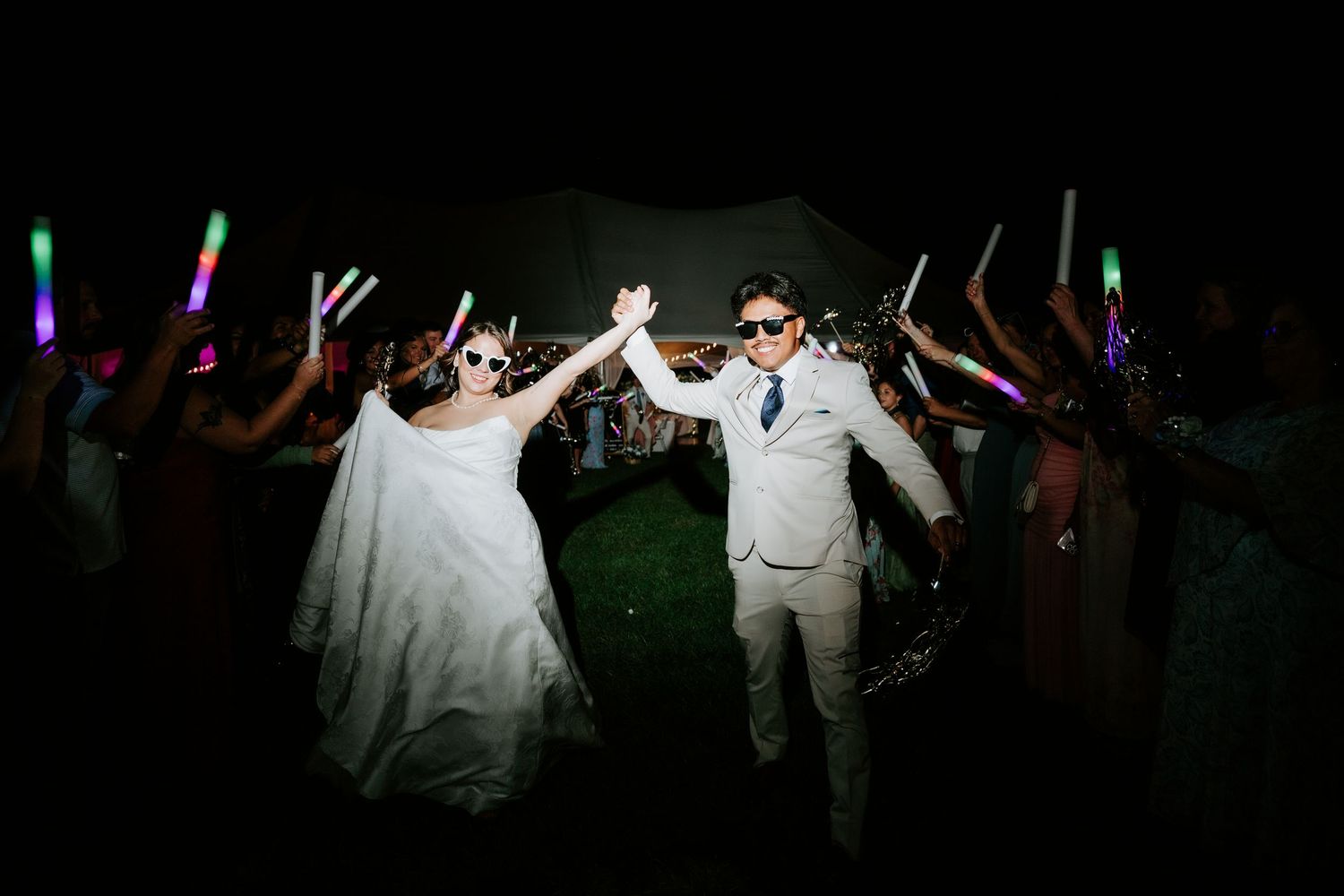 Bride and groom exit wedding reception through glowing foam stick tunnel with guests cheering at night.
