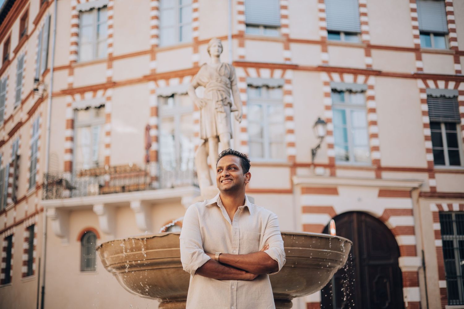 Man in white shirt stands arms crossed by ornate fountain with classical statue and historic building backdrop.
