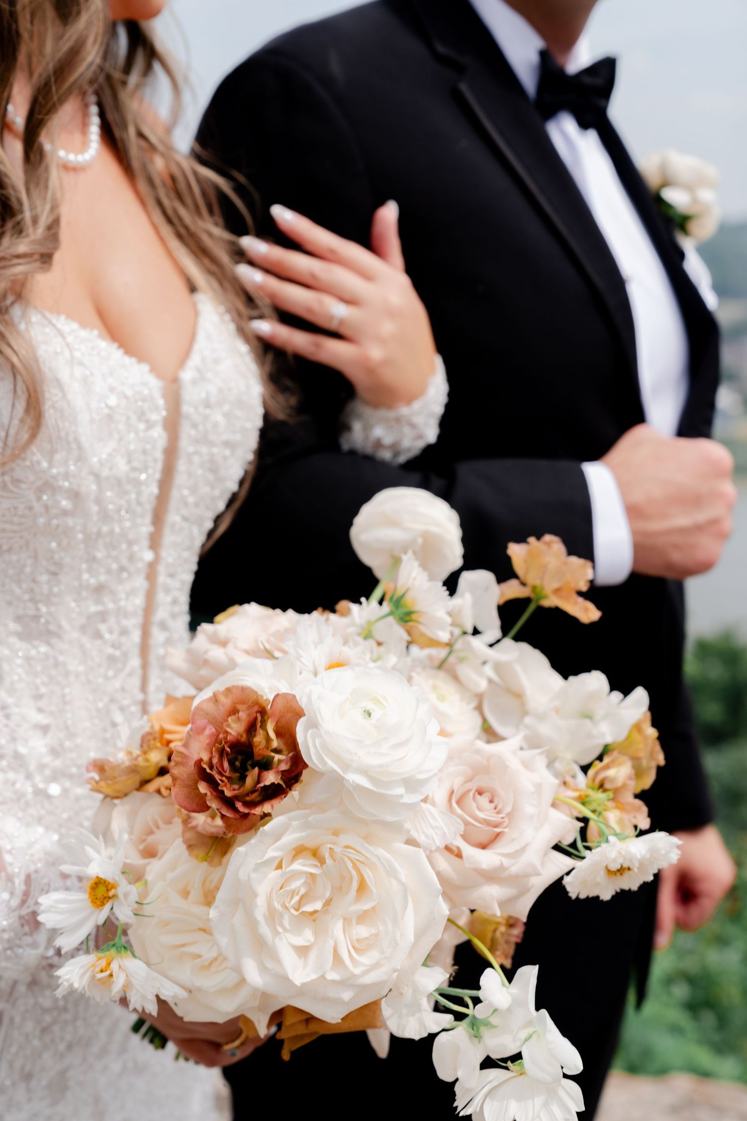 Bride holding a lush wedding bouquet of white and blush roses with rust blooms, standing beside groom in black tuxedo.