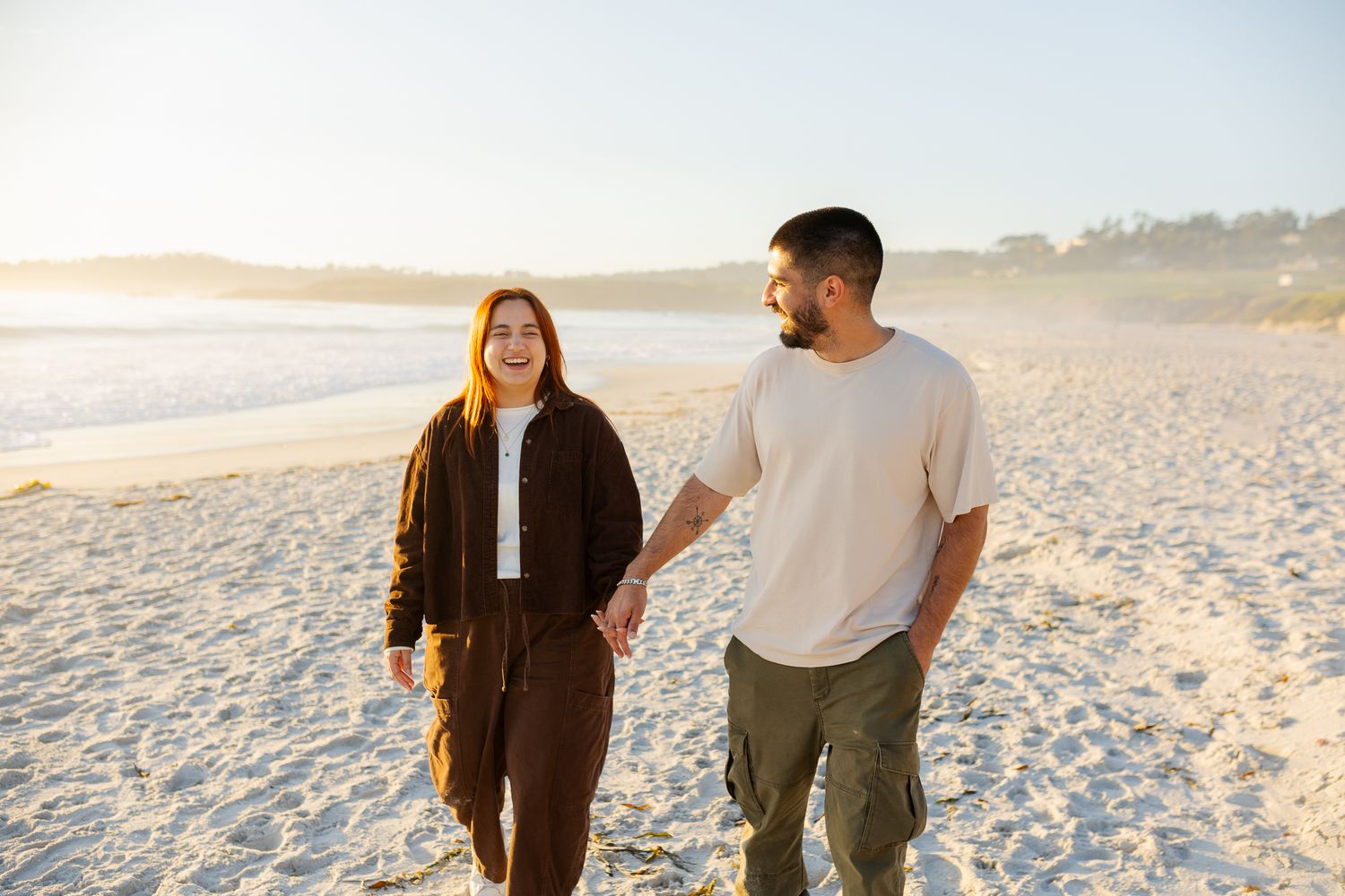 A happy couple holds hands while walking on a sunny beach at golden hour.
