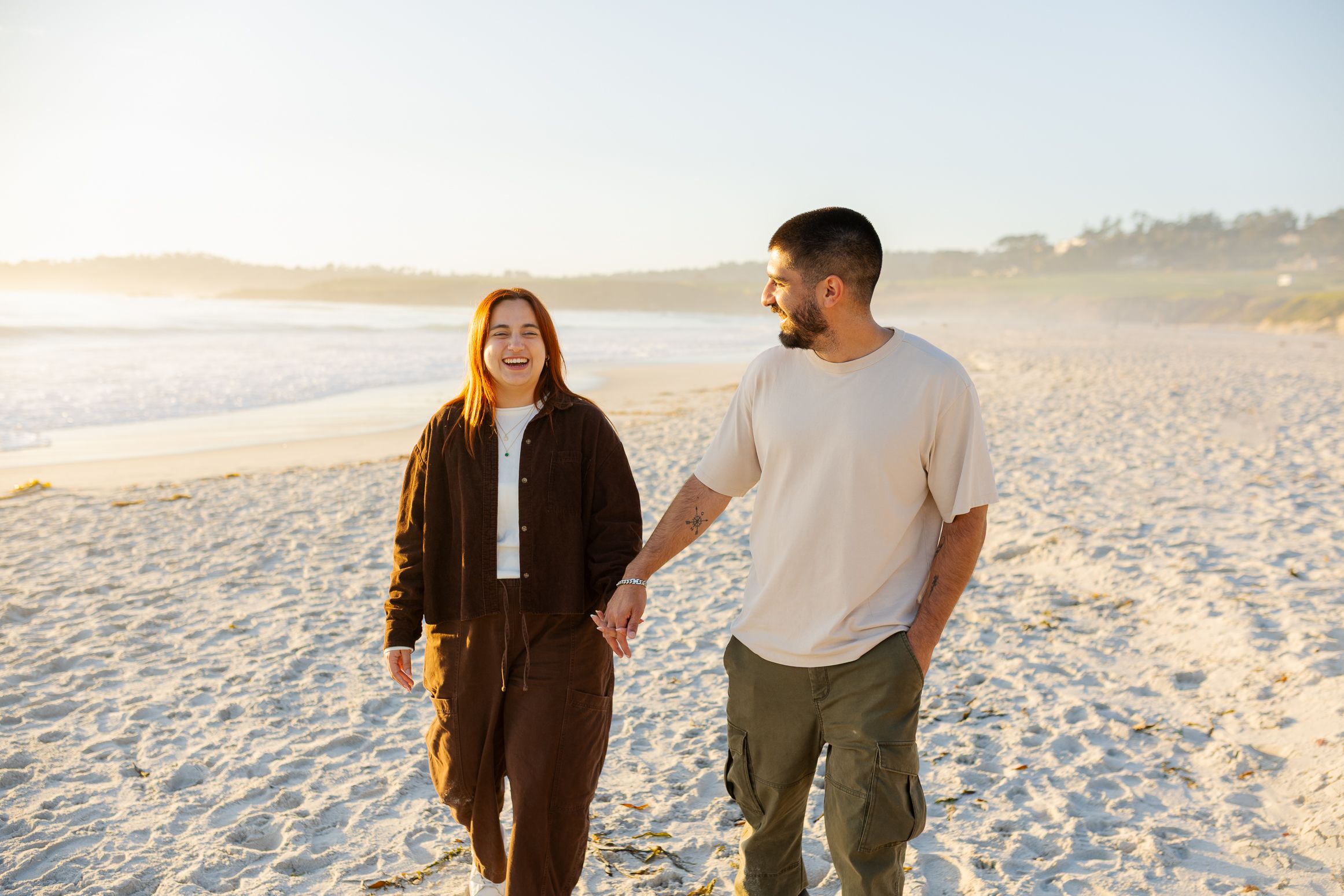 A happy couple holds hands while walking on a sunny beach at golden hour.