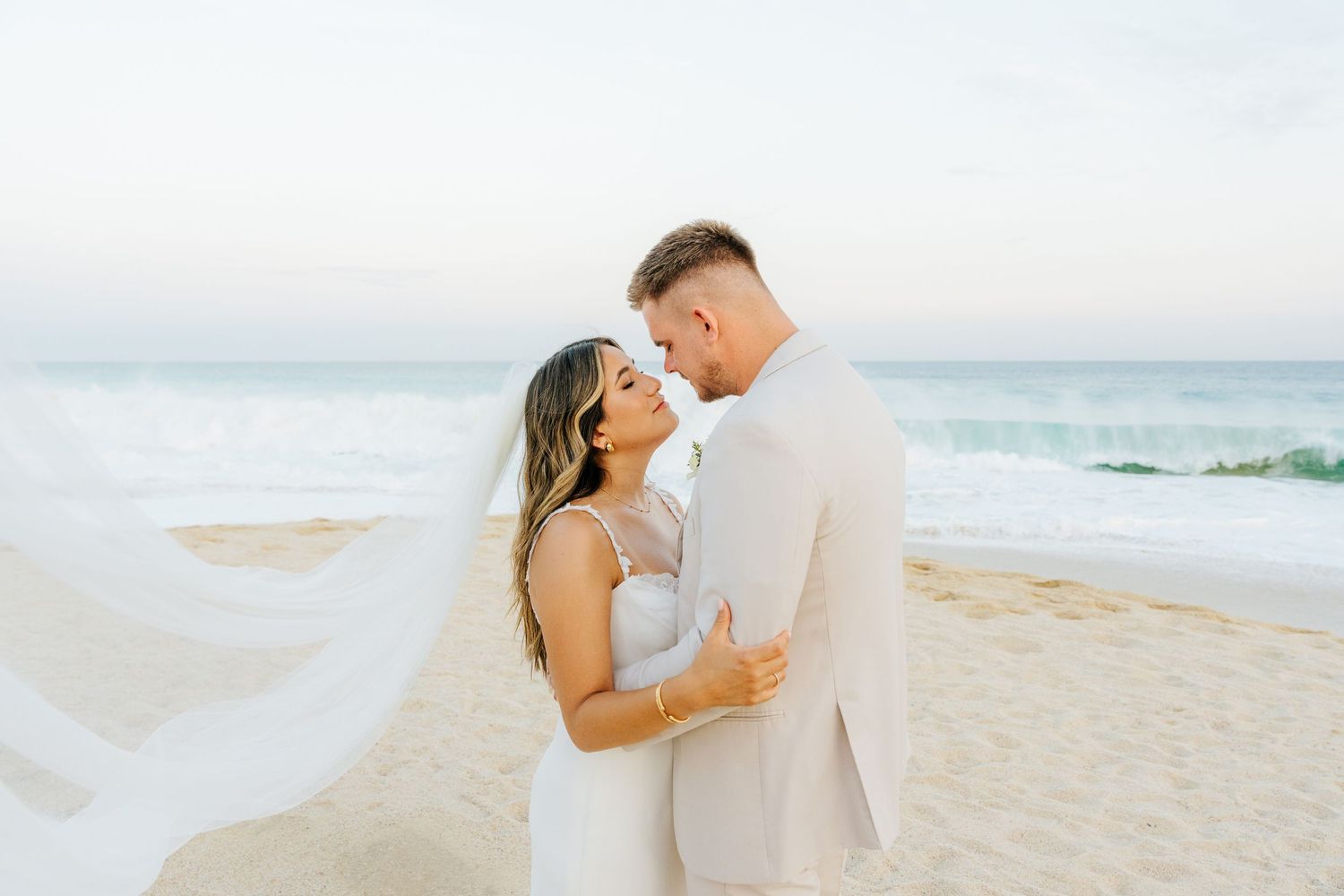 Bride in a white gown and groom in a cream suit share an intimate moment on a sandy beach as her veil blows in the ocean breeze.