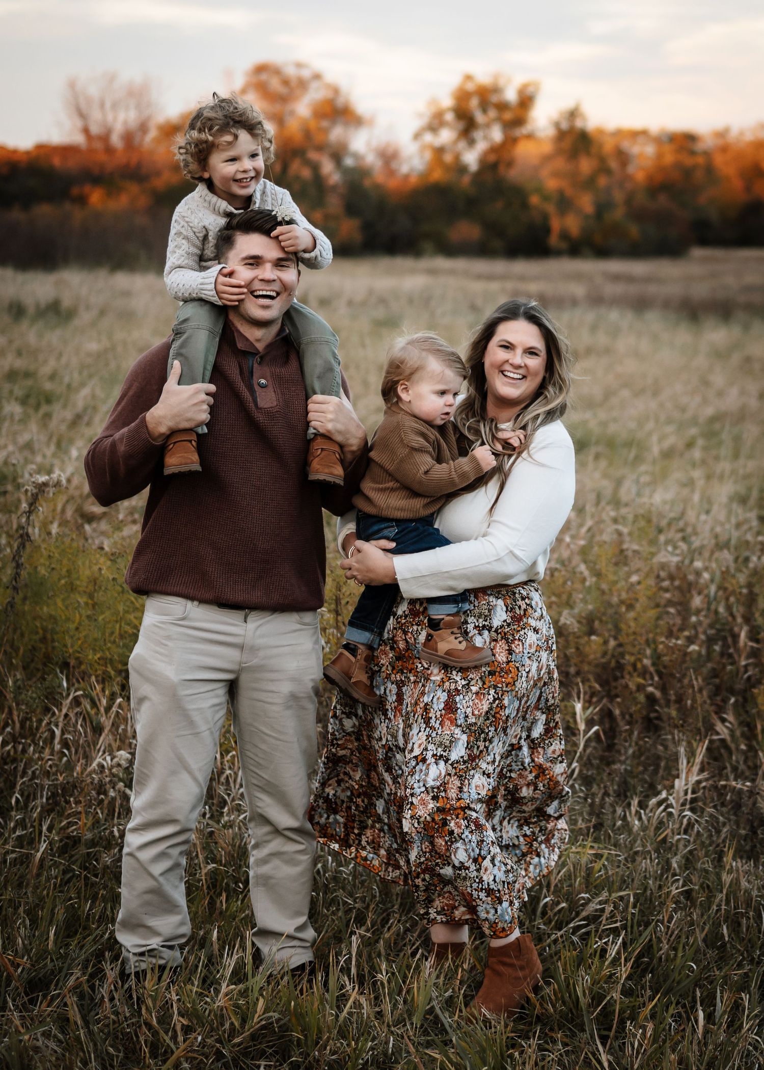 Happy family of four posing in autumn field at sunset, father with toddler on shoulders, mother holding baby.