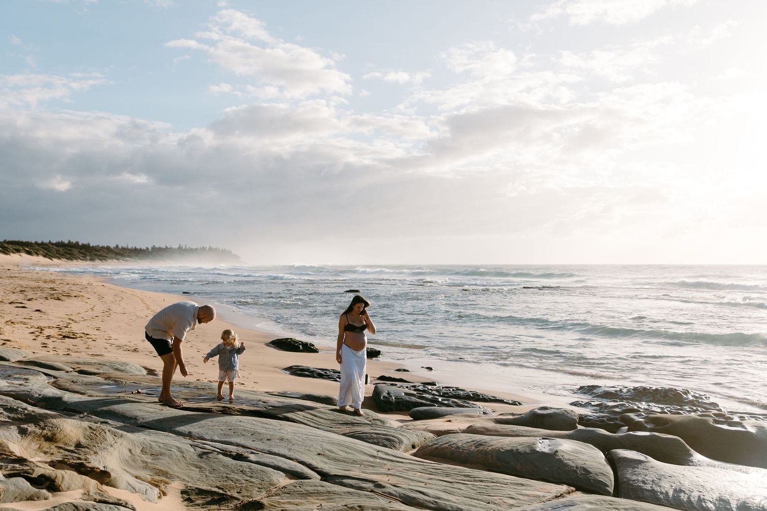 Family enjoying a sunny beach day on rocky shores with ocean waves and sandy coastline in the background.