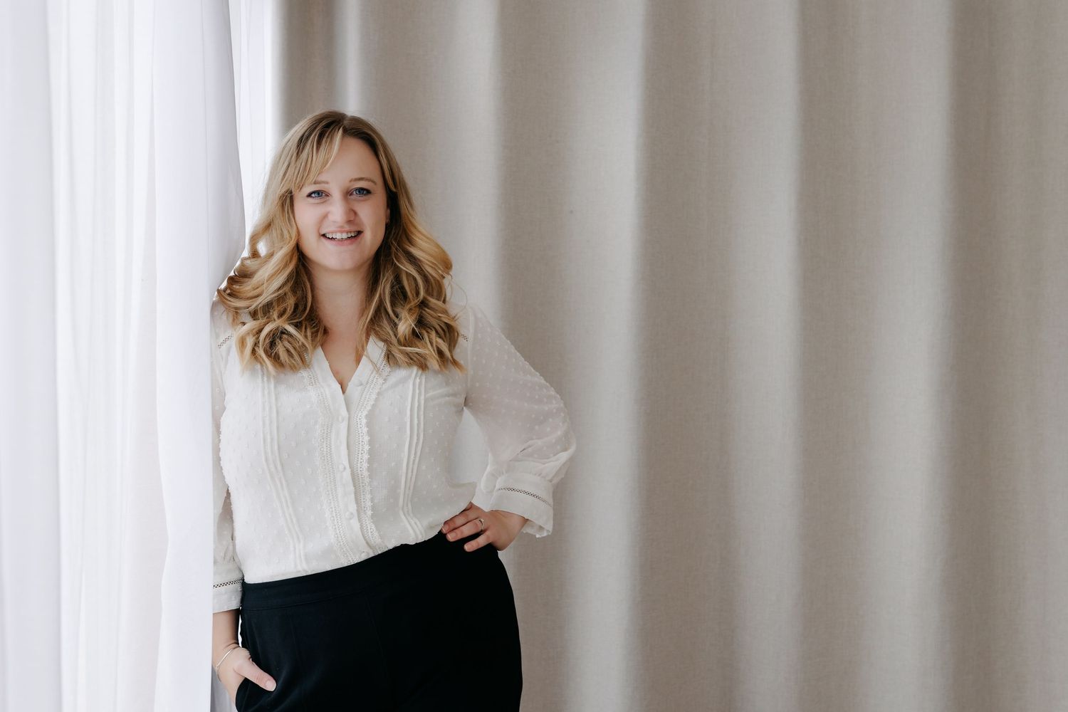 Smiling blonde woman in white blouse and black pants posing confidently against soft curtain backdrop.
