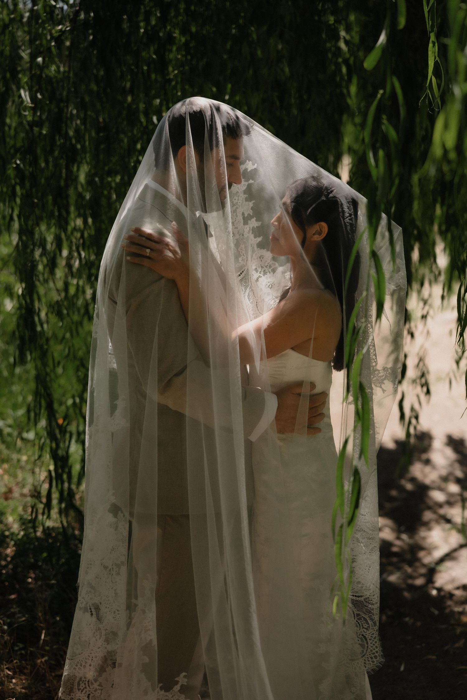 Black and white photo of bride and groom sharing an intimate moment under flowing wedding veil in outdoor field.