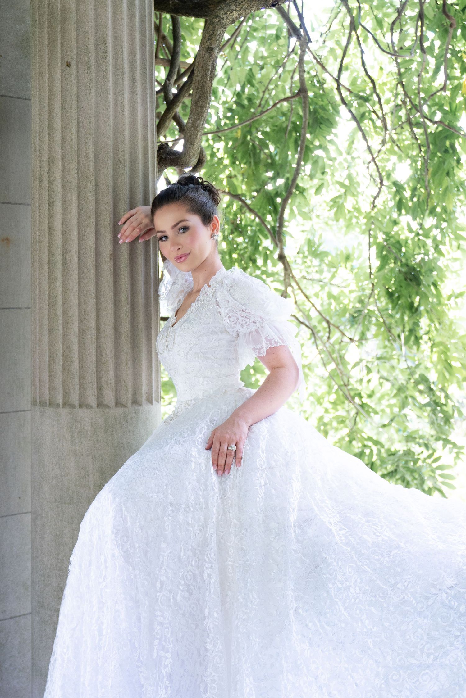 Bride in flowing white lace ball gown posing beside stone column surrounded by green trees.