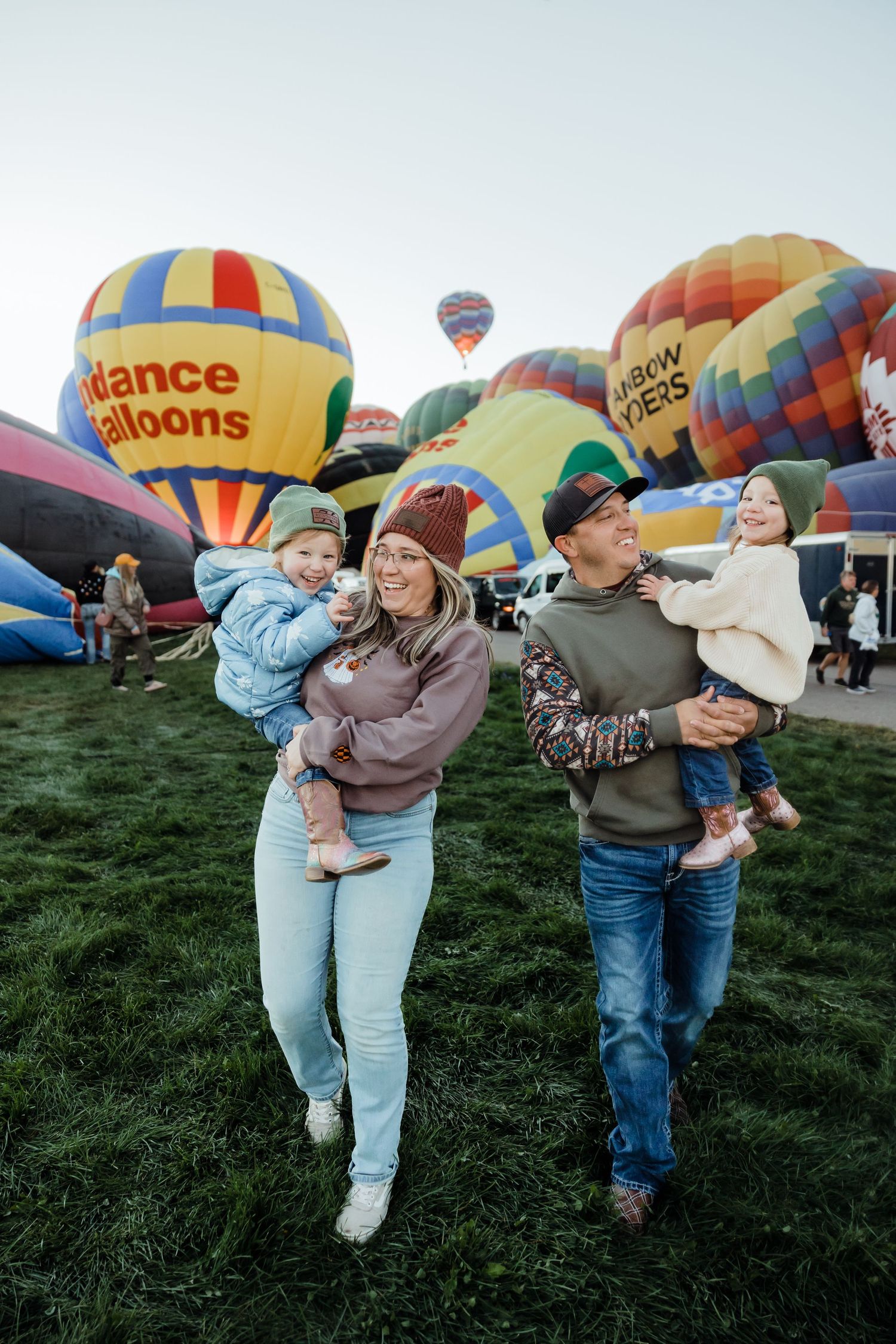 A happy family of four poses at a colorful hot air balloon festival on a green field at dusk.
