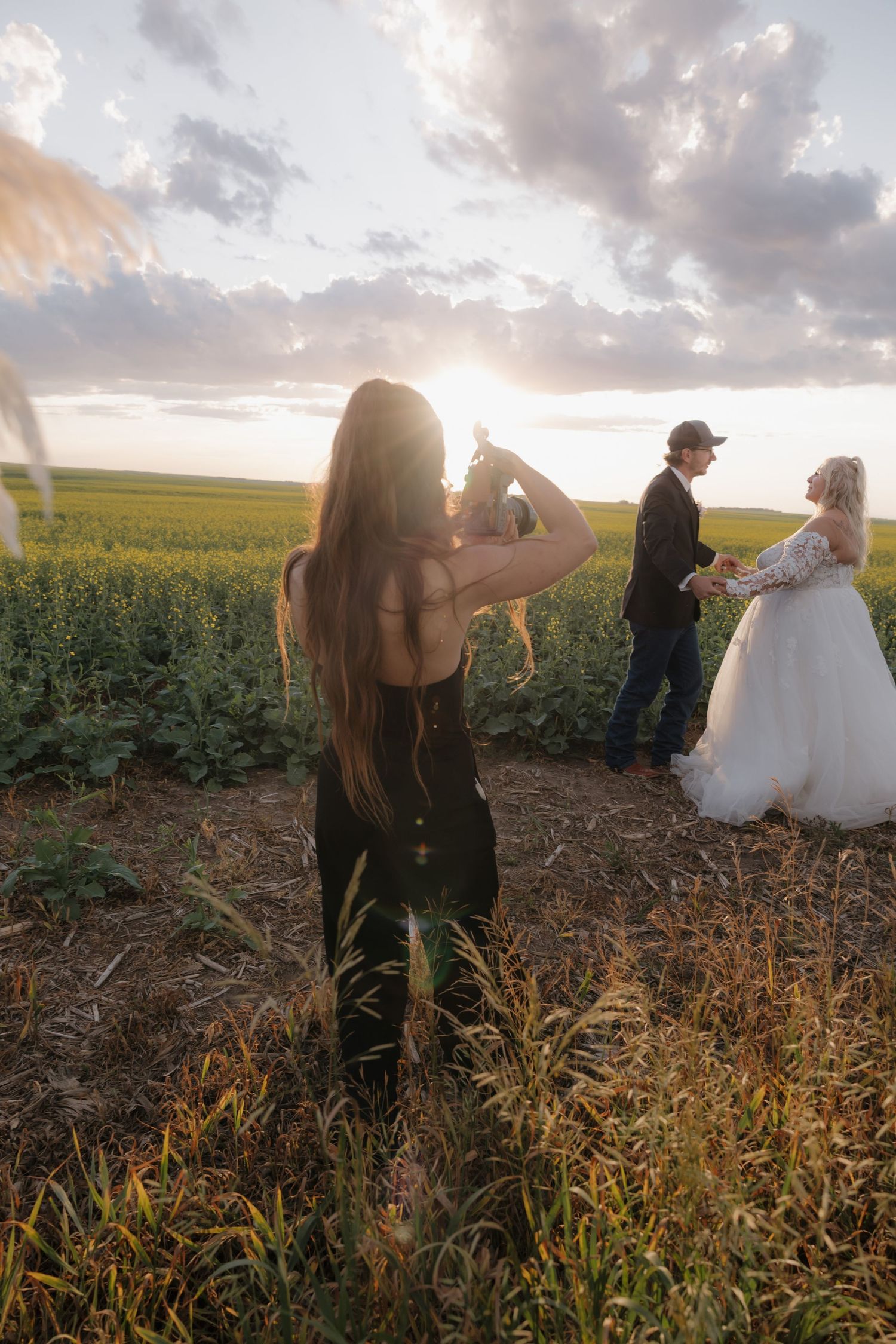 Couple embraces in a golden sunset field while photographer captures romantic moments during their wedding shoot. Garrison, ND
