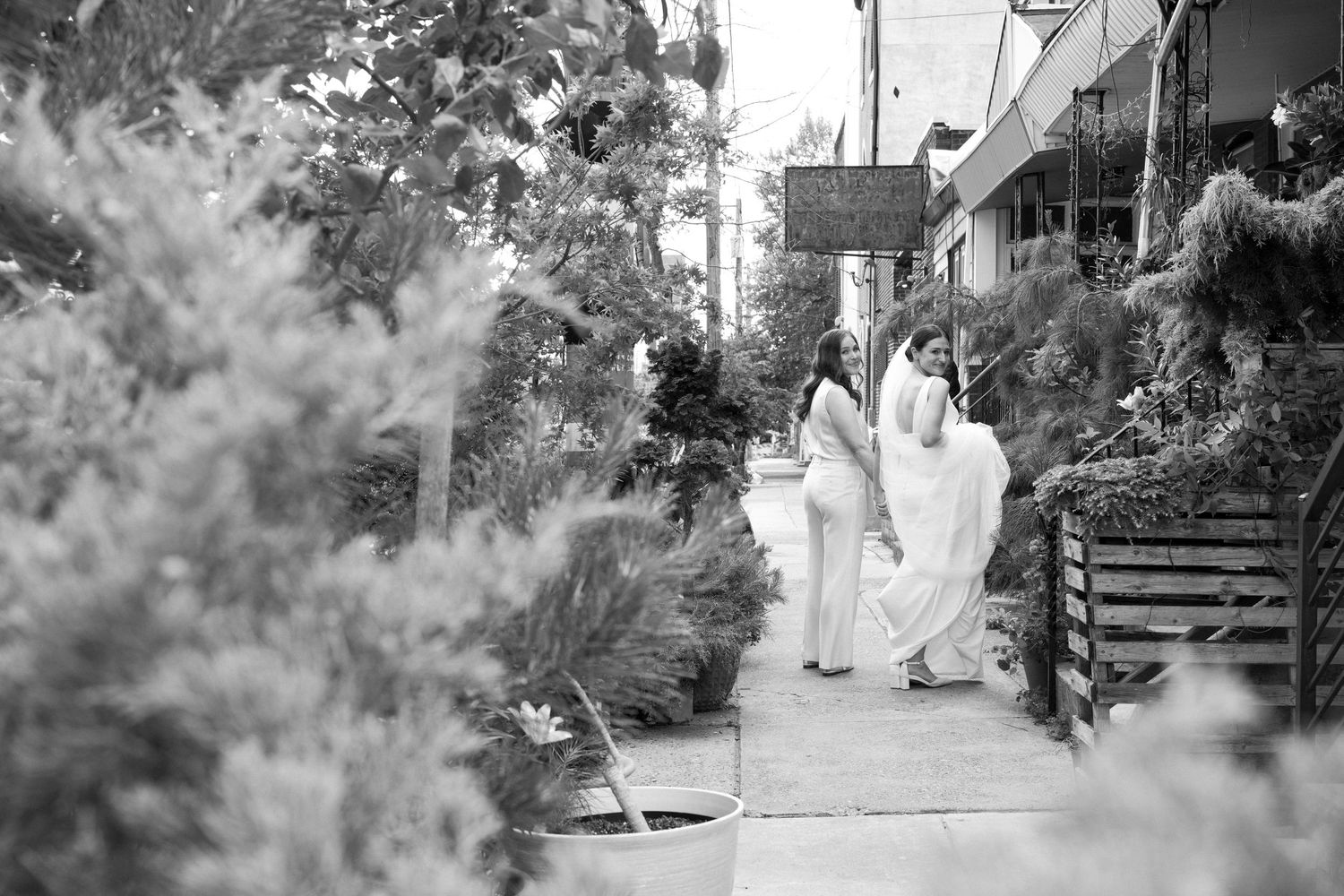 Black and white photo of women in elegant white wedding attire, one wearing a sleek pantsuit and one in backless gown with buttons.