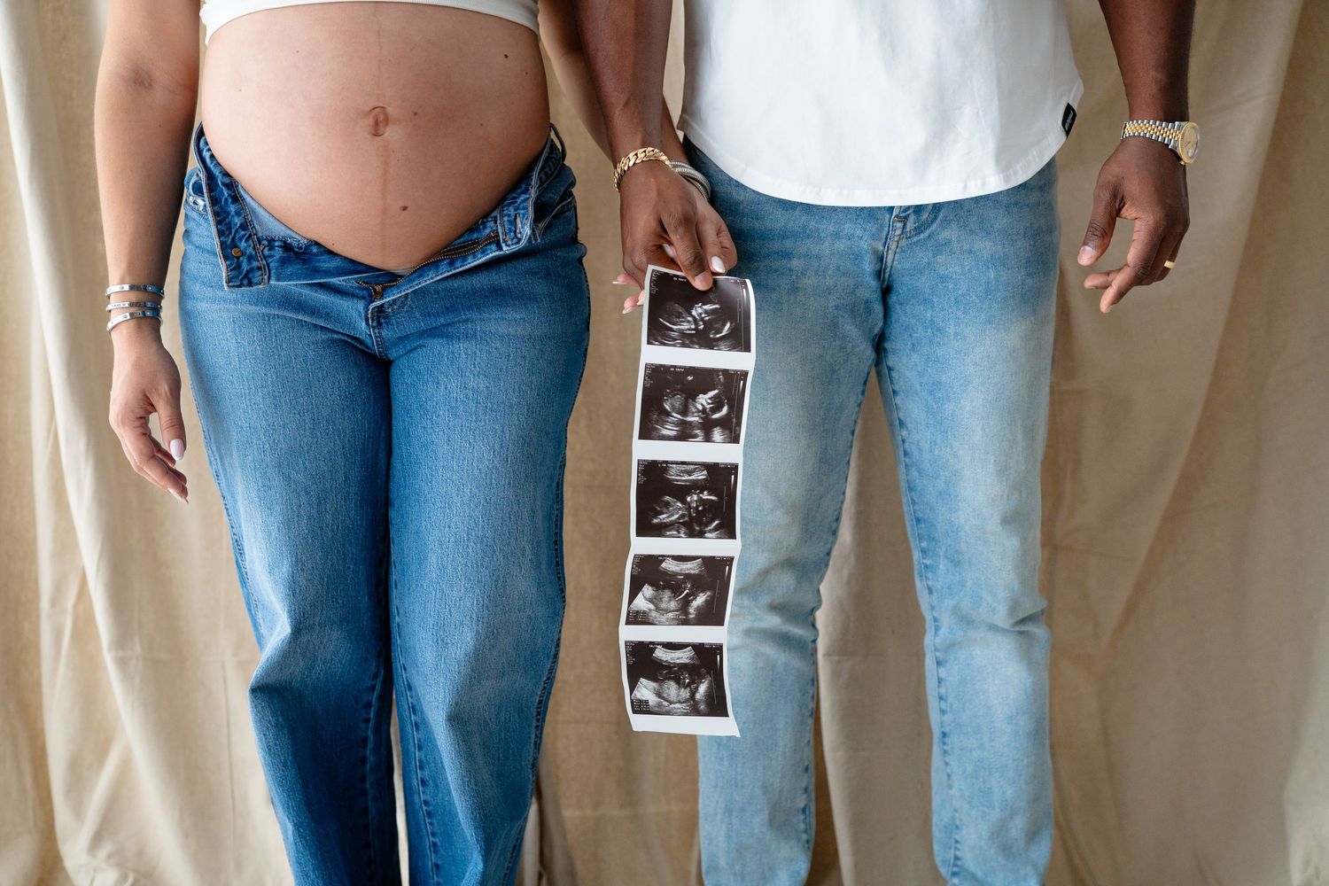 Expecting couple stands together holding ultrasound photos while showing pregnant belly in denim jeans.
