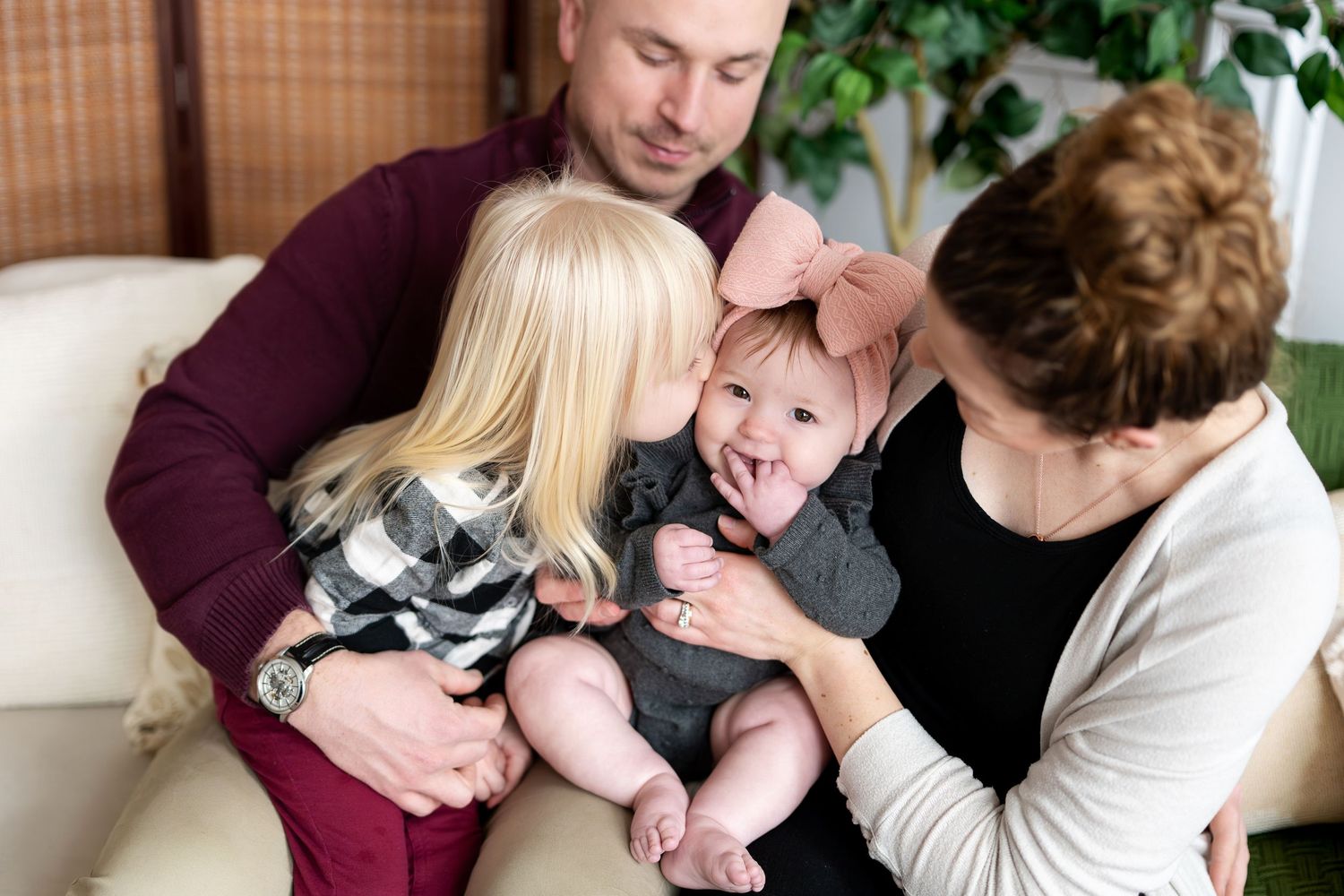 Family with bald father, blonde toddler girl, and grandmother holding a baby in gray outfit indoors.
