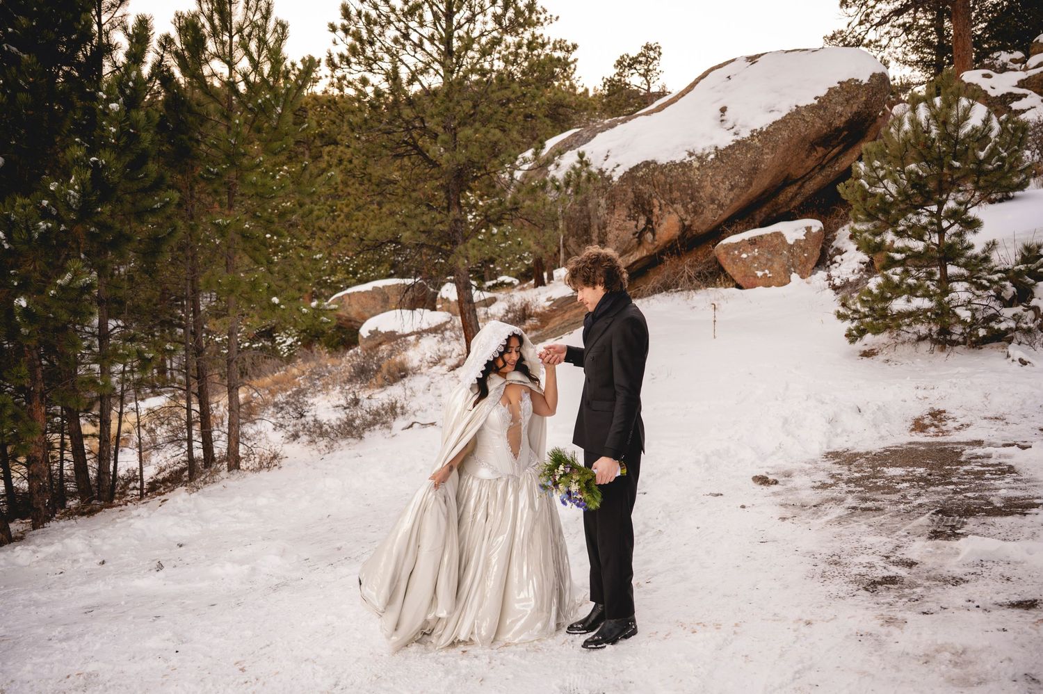 A couple in formal attire poses together in a snowy winter landscape surrounded by evergreen trees.