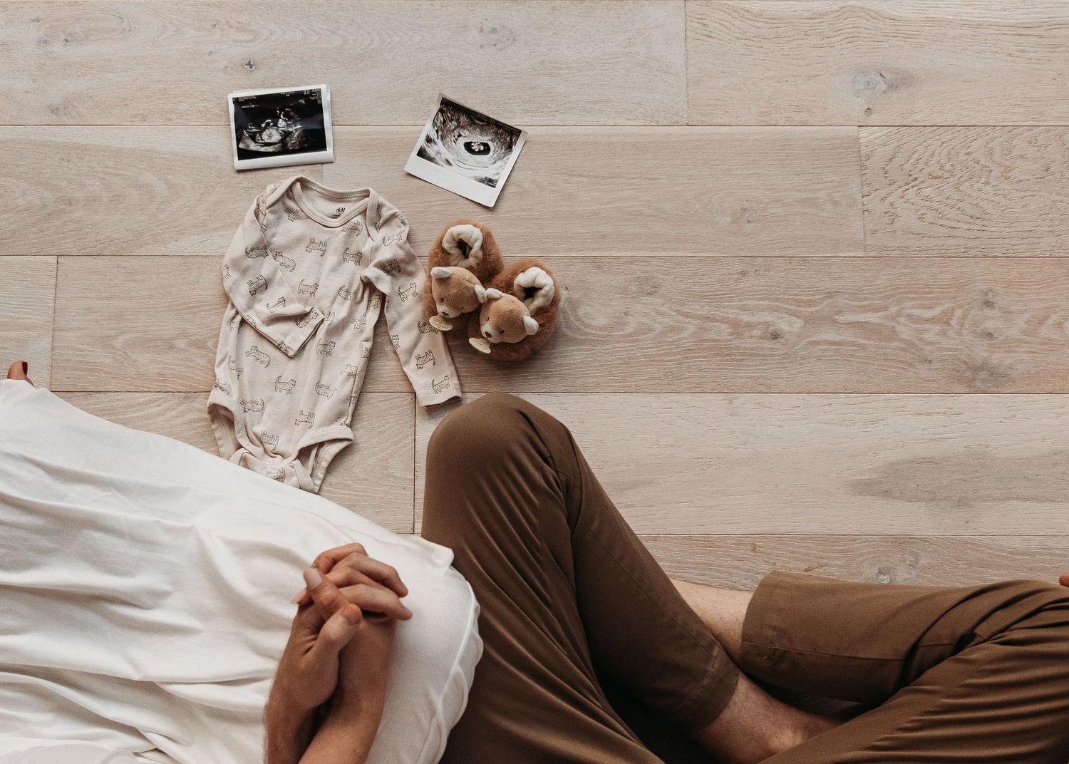 Sonogram ultrasound photos displayed on wooden floor beside baby onesie and teddy bear in pregnancy announcement style.