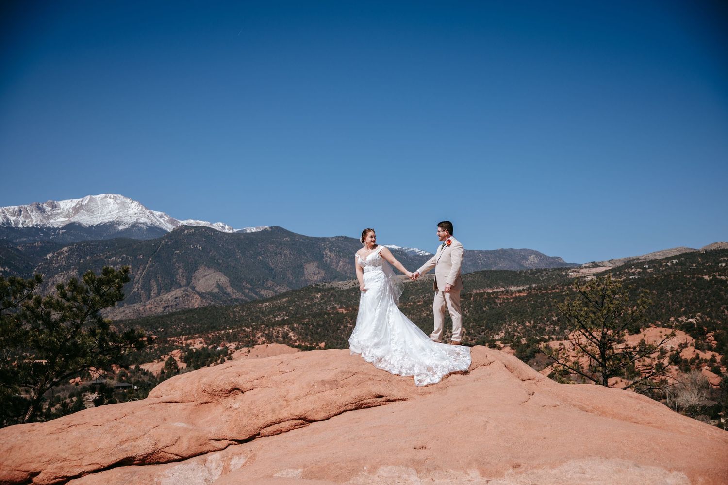 A couple in wedding attire stands on red rocks with Pikes Peak mountain vista in Colorado Springs.