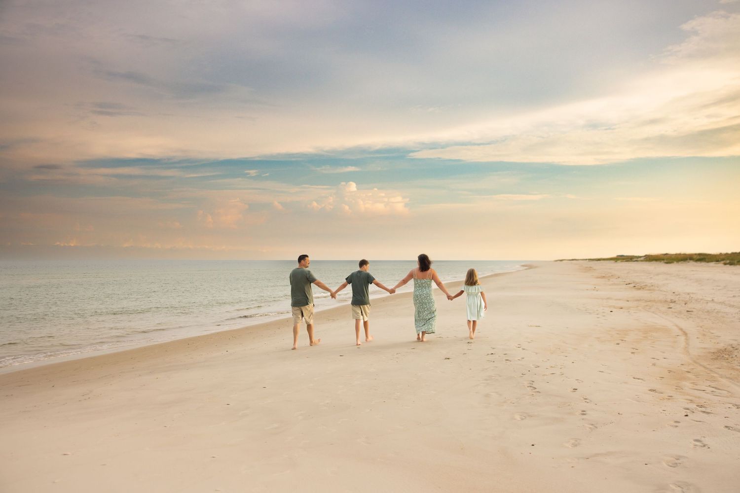 Family silhouettes hold hands and skip together on empty beach at sunset with peaceful ocean view. St. George Island, Florida.