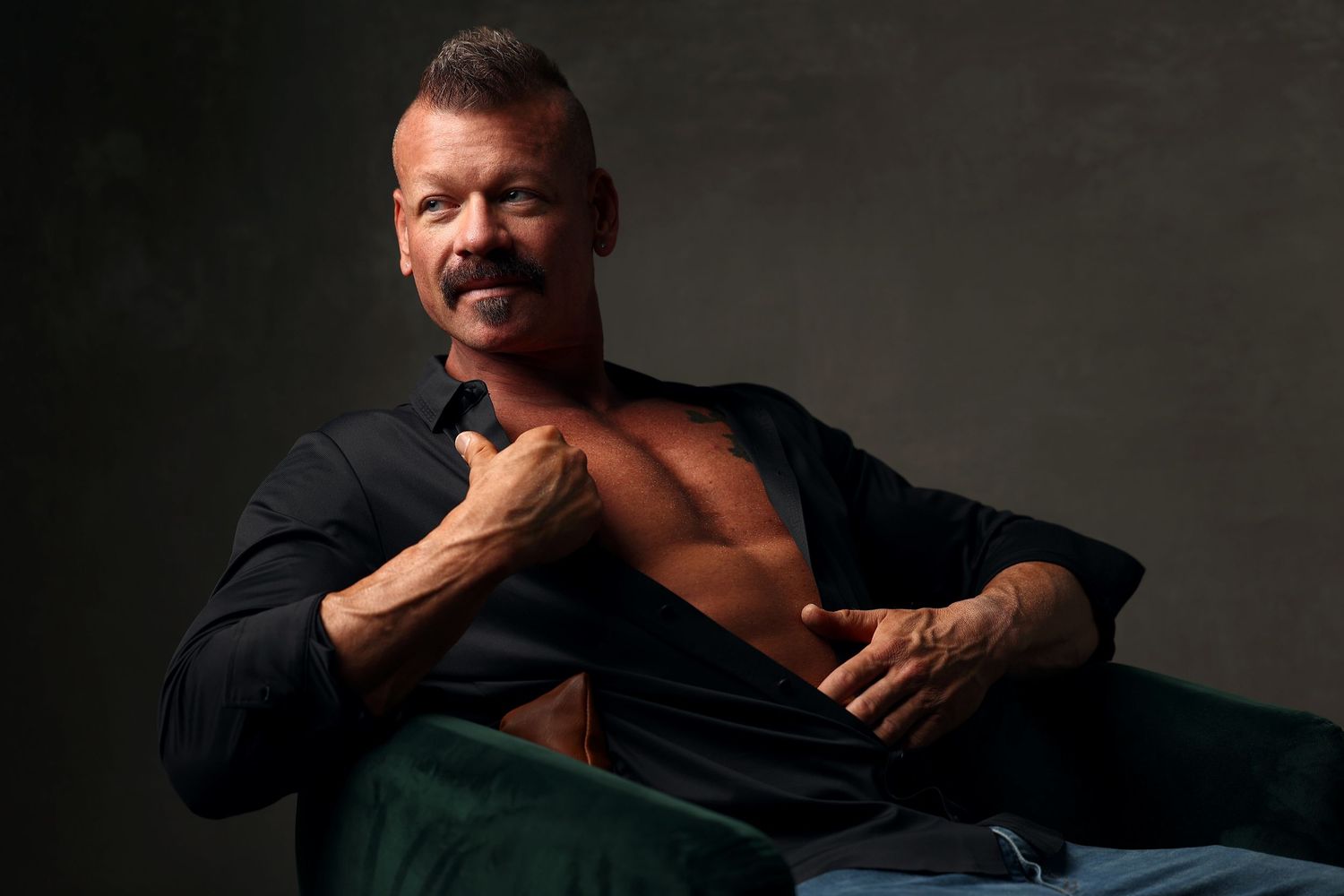 Male fitness competitor sitting in a chair, shirt half unbuttoned, relaxed pose highlighting chest and abs, post-competition boudoir photosh