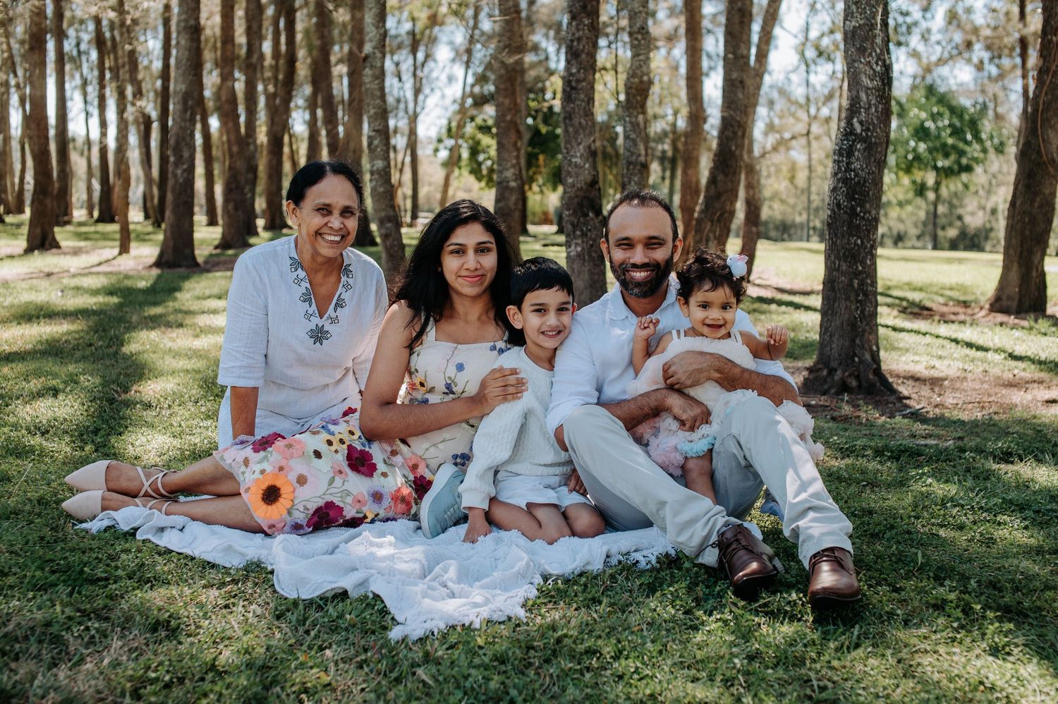 A family enjoys a picnic together on a white blanket in a sunny wooded area.