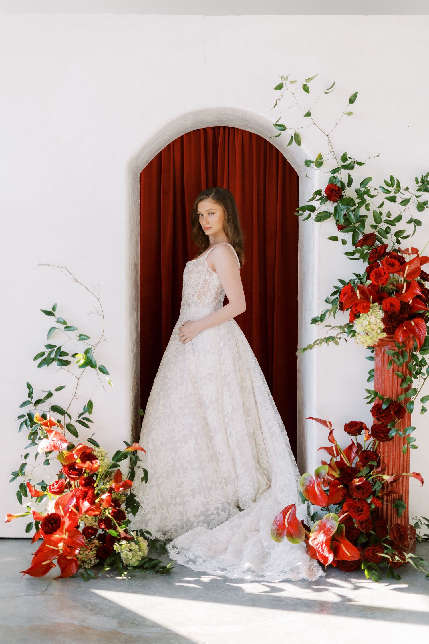 A bride wearing an elegant white wedding gown and veil with delicate floral appliques against a red curtain backdrop.