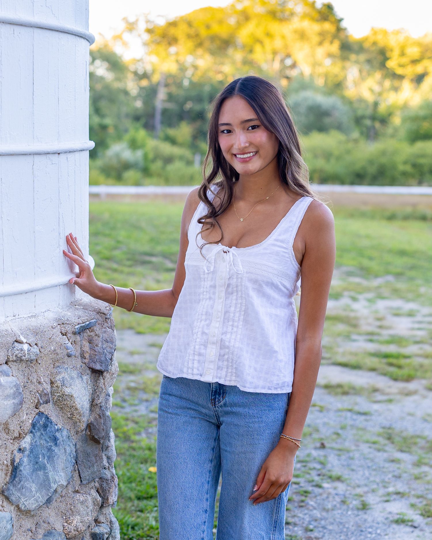 High school senior in a white sleeveless top and blue jeans poses outdoors against a silo with a fieldstone foundation at a farm in Sudbury