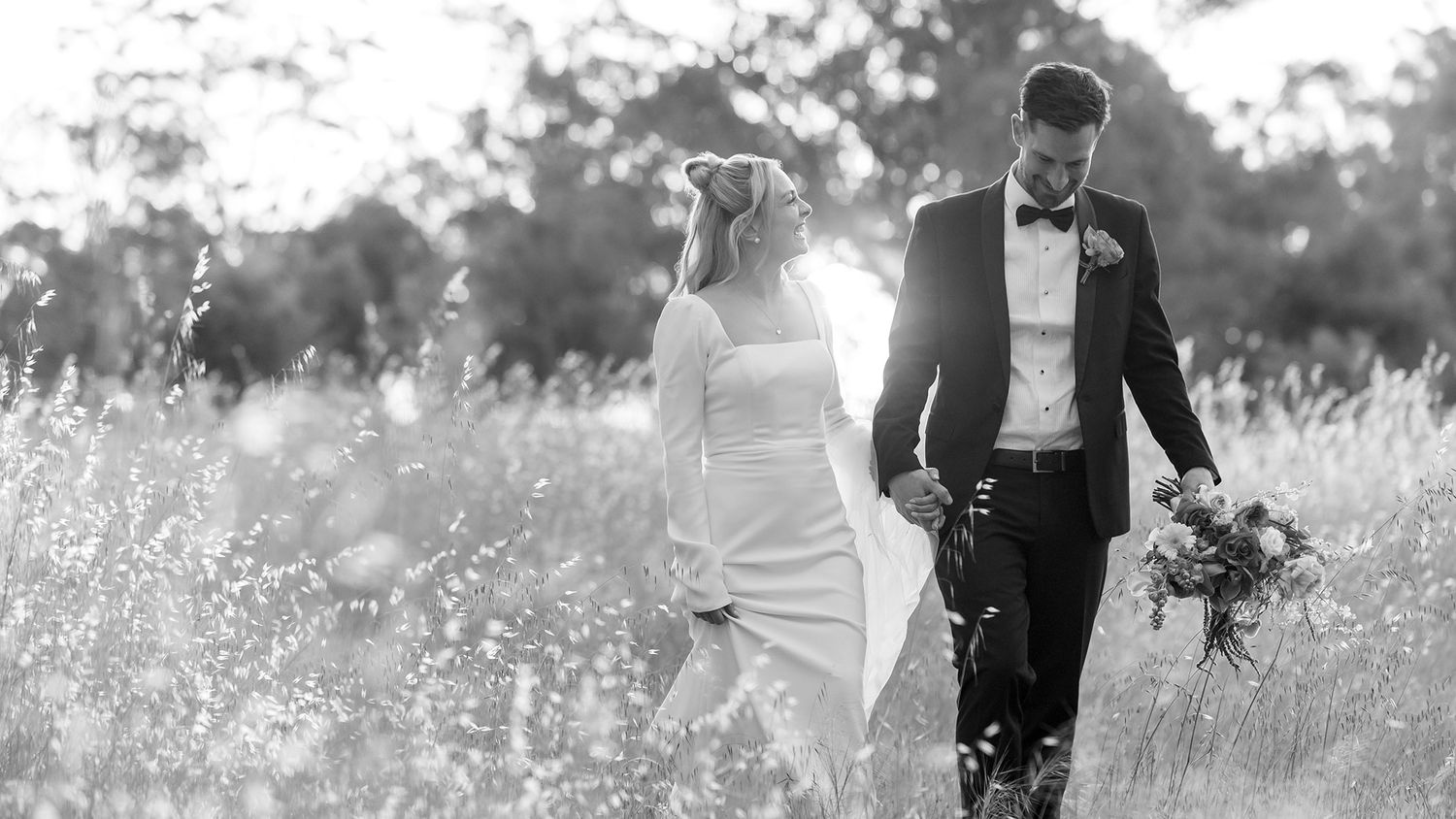 A black and white image of a Bride and Groom walking in long grass holding hands with the sun setting in the background